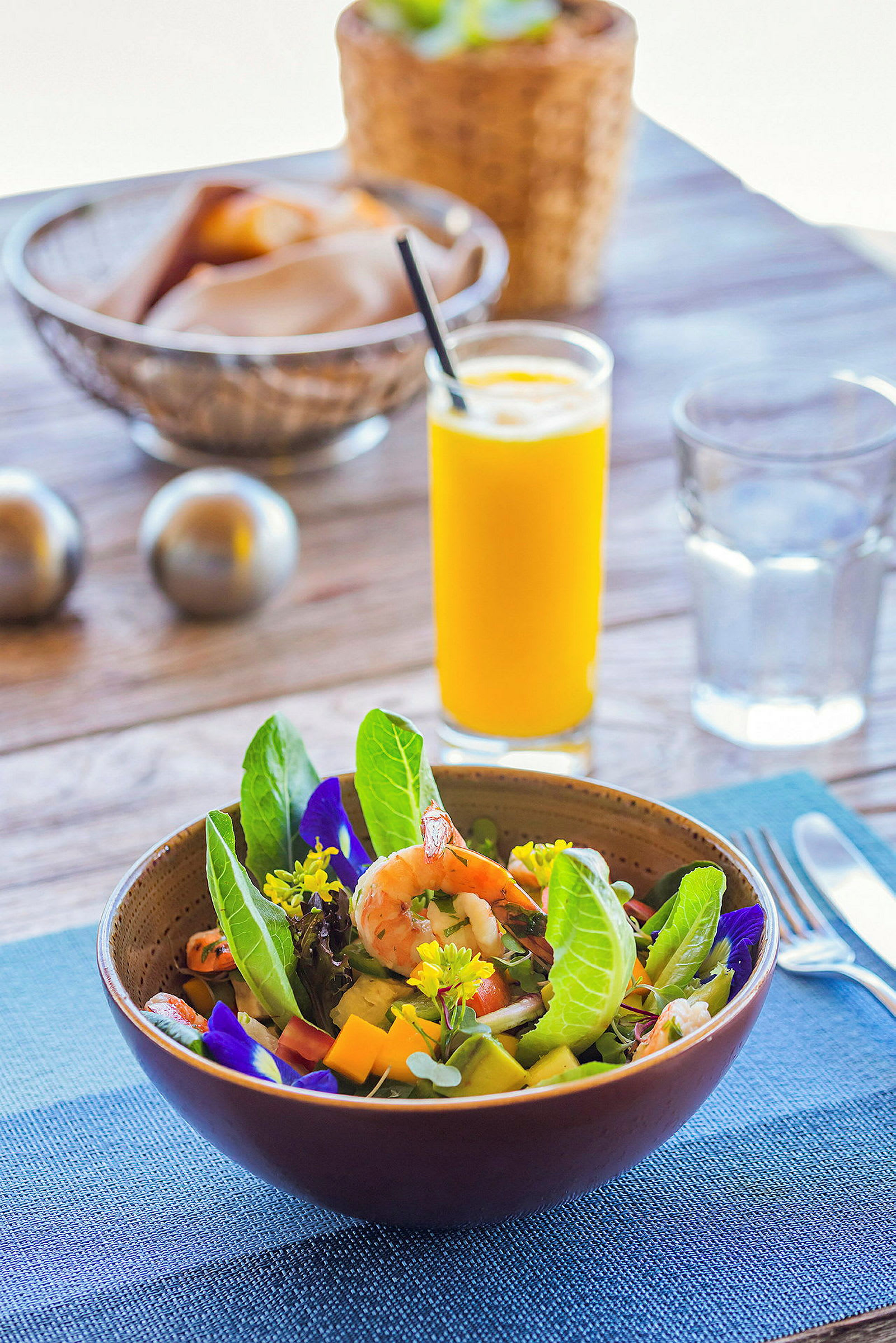 En salat i en skål med rejer, grønne blade og blomster. Et glas appelsinjuice og brød i baggrunden. (AI-genereret tekst)