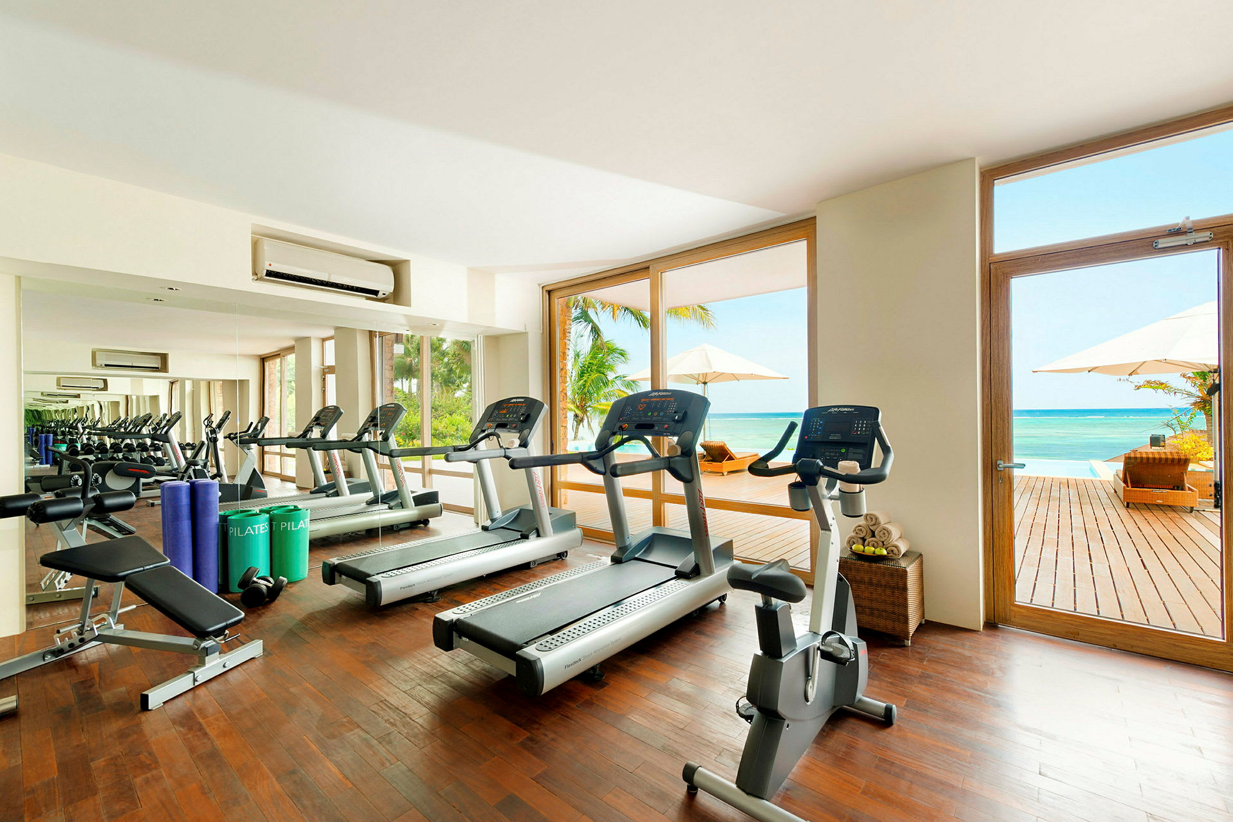 Et fitnesscenter med træningsmaskiner og spejlvæg, store vinduer åbner op mod en terrasse med udsigt over havet. (AI-genereret tekst)