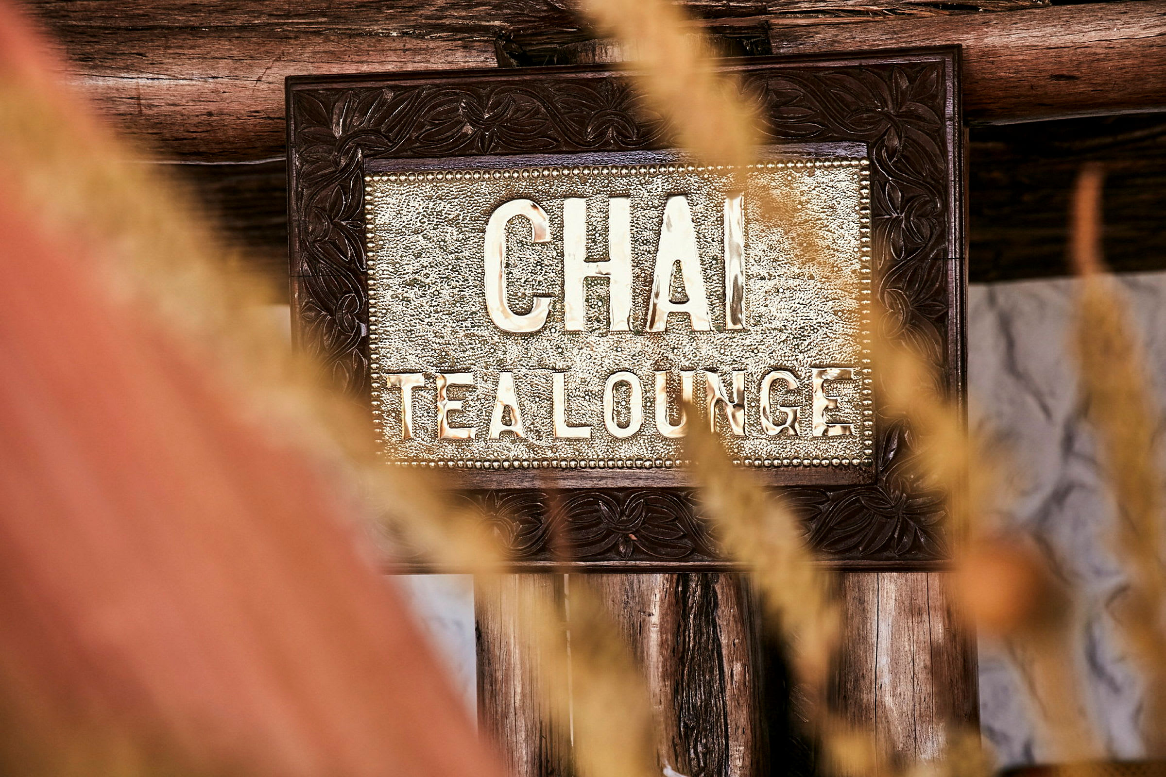 Et nærbillede af et skilt med teksten 'Chai Tea Lounge', omgivet af trædekorationer og visnende græs i forgrunden. (AI-genereret tekst)