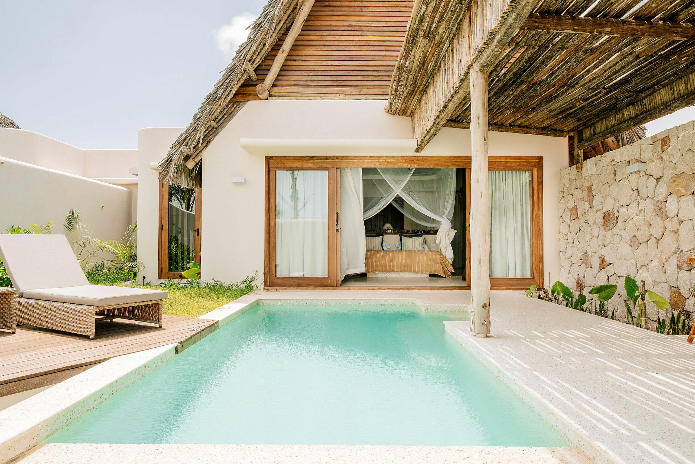En villa med stråtag og store vinduer, foran en privat pool med en liggestol på en træplatform. (AI-genereret tekst)
