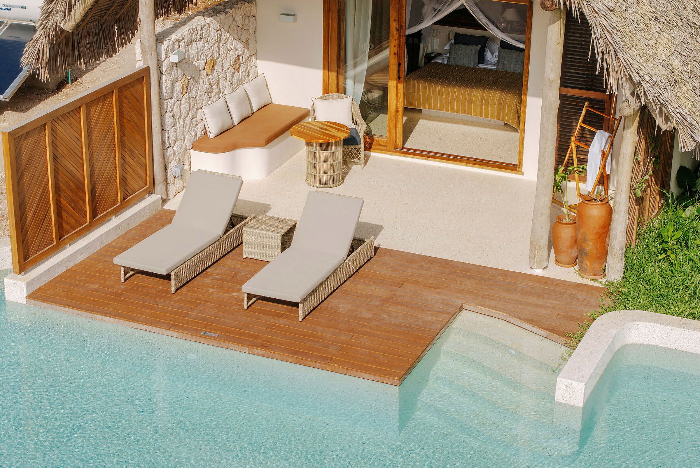 En træterrasse ved en pool med to liggestole og et loungeområde. Bag terrasseområdet er der en villa med glasdøre og stråtag. (AI-genereret tekst)