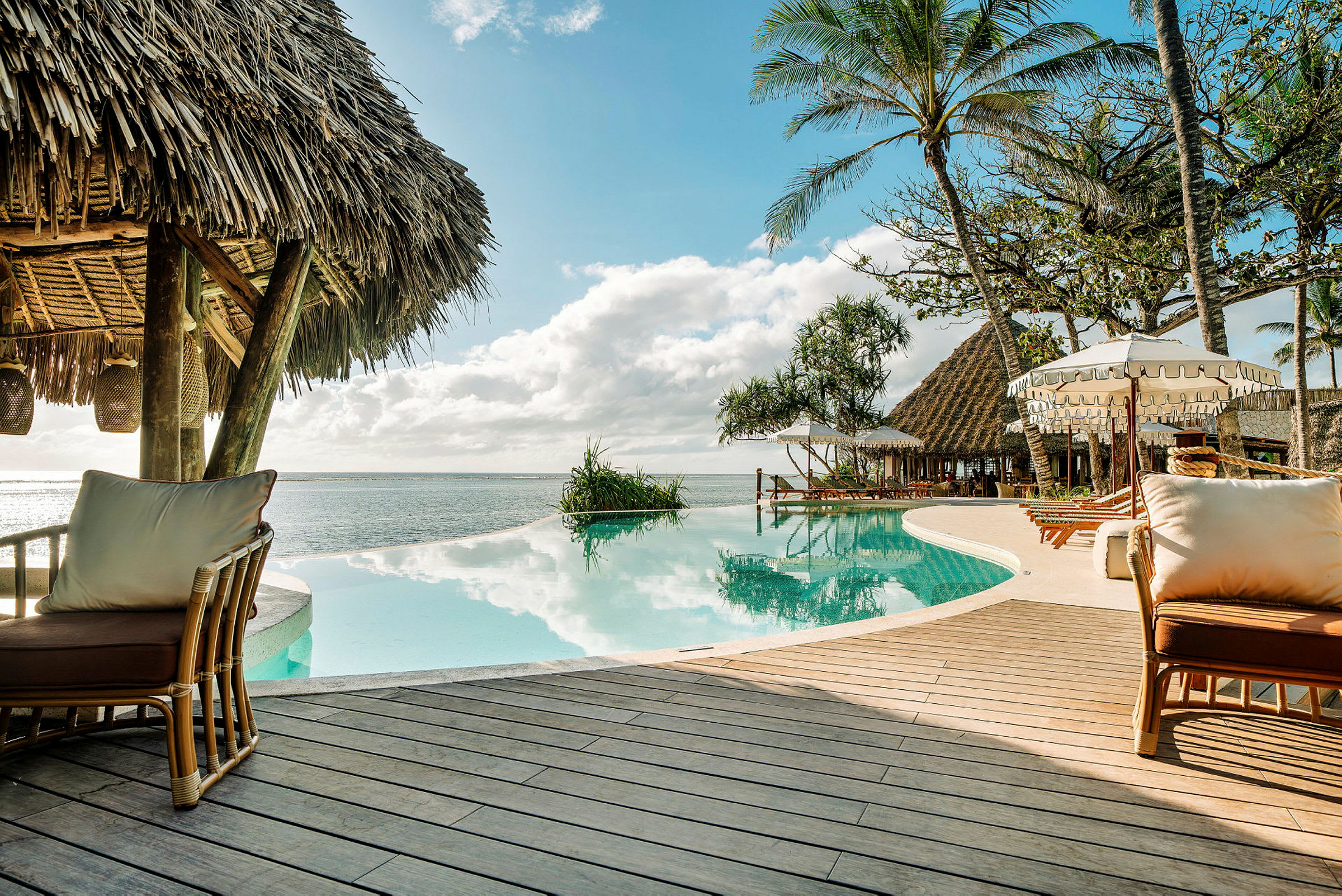 Billede af en infinity-pool ved en strand med stråtag, liggestole og parasoller i en tropisk miljø. (AI-genereret tekst)