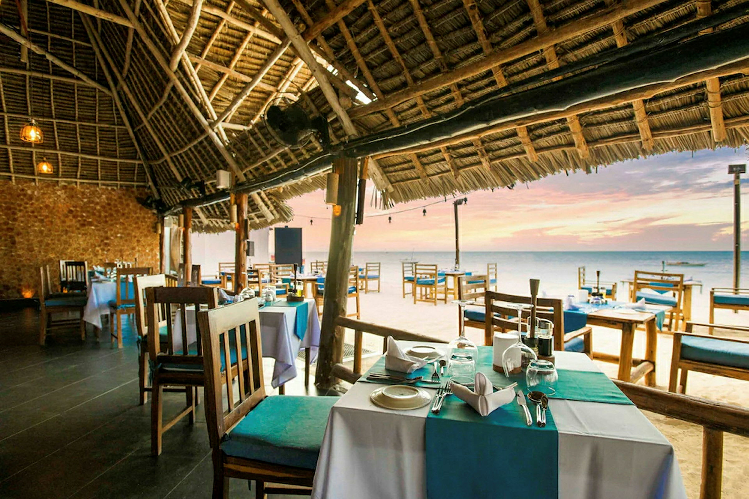 Billede af en strandrestaurant med borde og stole under et stråtag. Havet og solnedgangen ses i baggrunden. (AI-genereret tekst)
