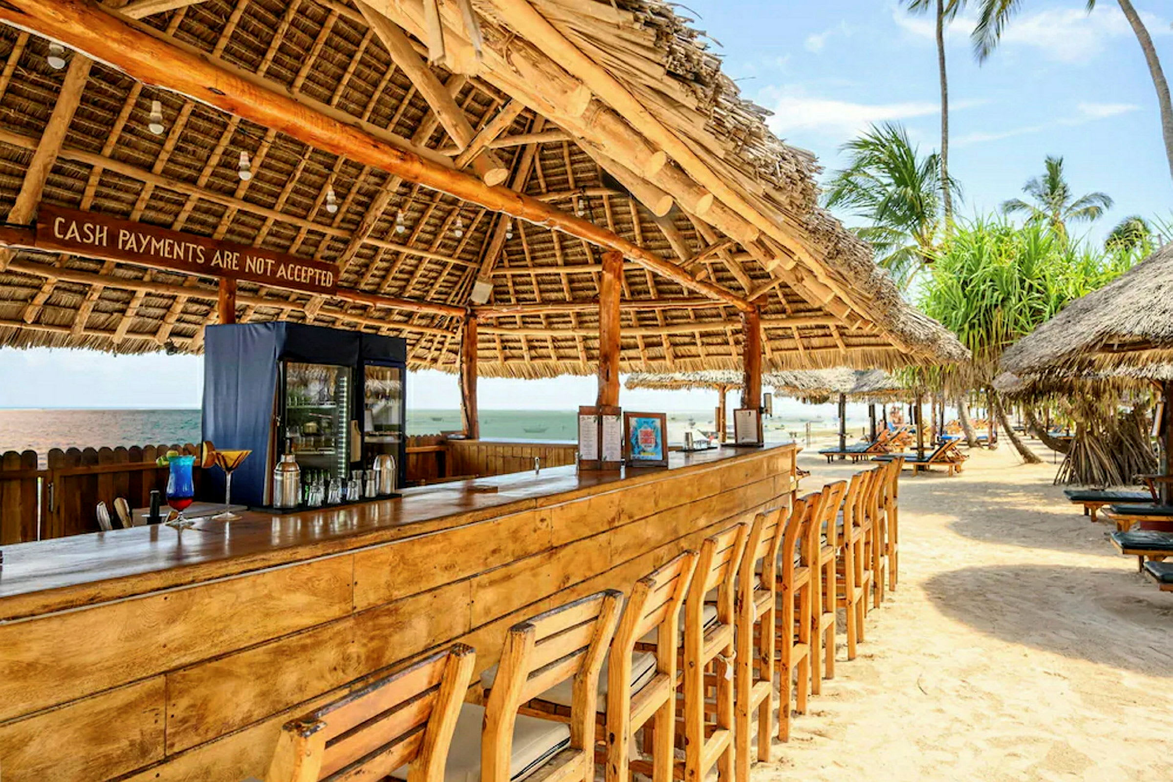 Strandbar med træbord og stole under tag af palmeblade. (AI-genereret tekst)