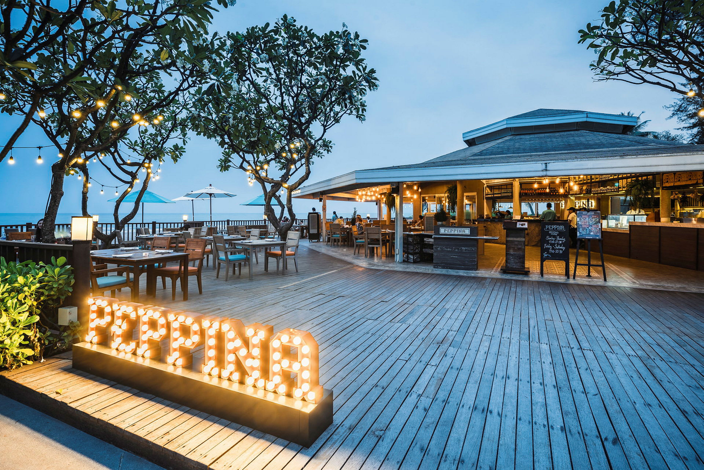 En restaurant med trædæk, borde og siddepladser udendørs nær havet. Skilt med oplyst teksten 'PEPPINA'. (AI-genereret tekst)