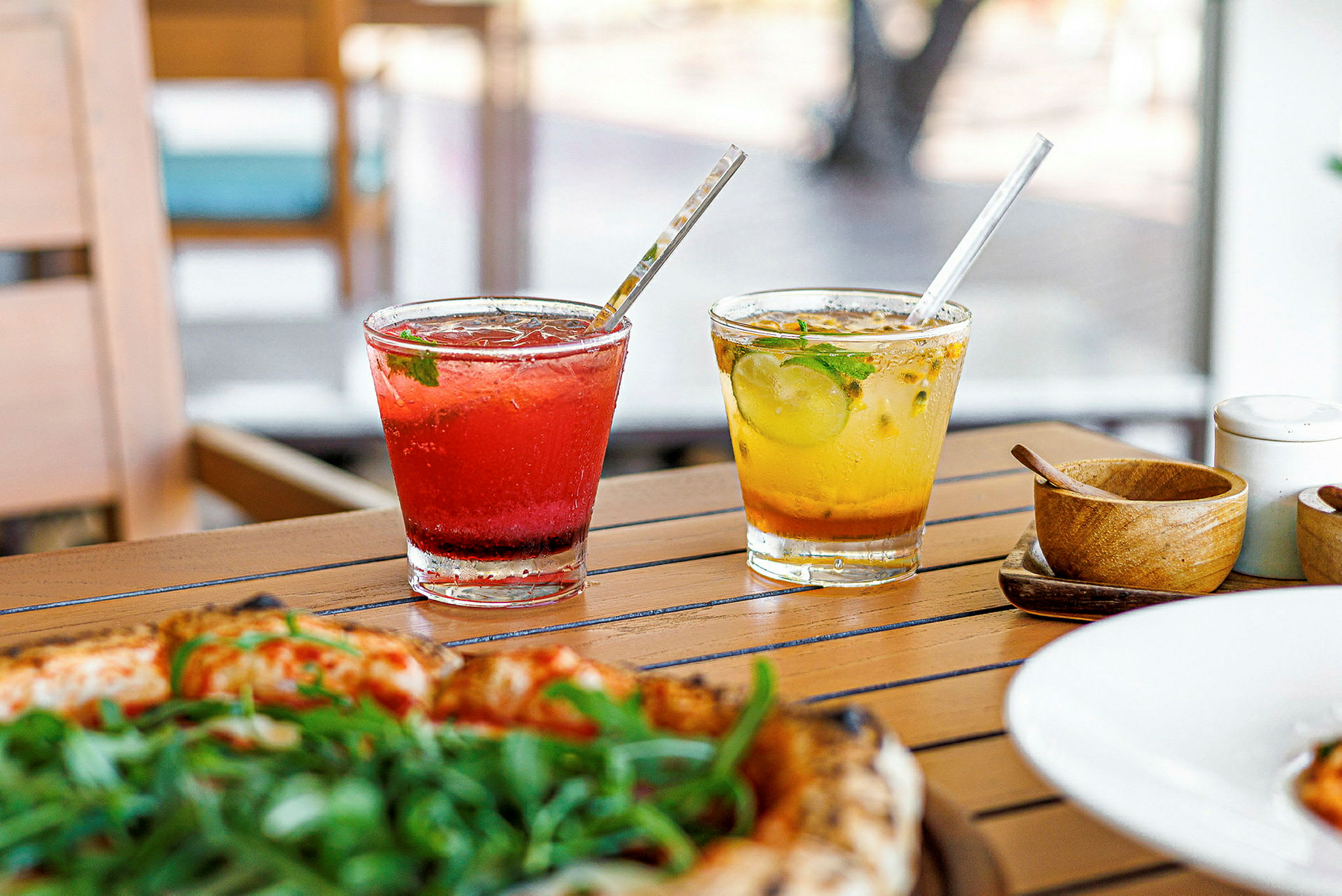 To glas med farverige drikke står på et træbord. Et glas indeholder rød drik, og det andet glas gul drik. Foran glassene ses en pizza med grøn topping. (AI-genereret tekst)