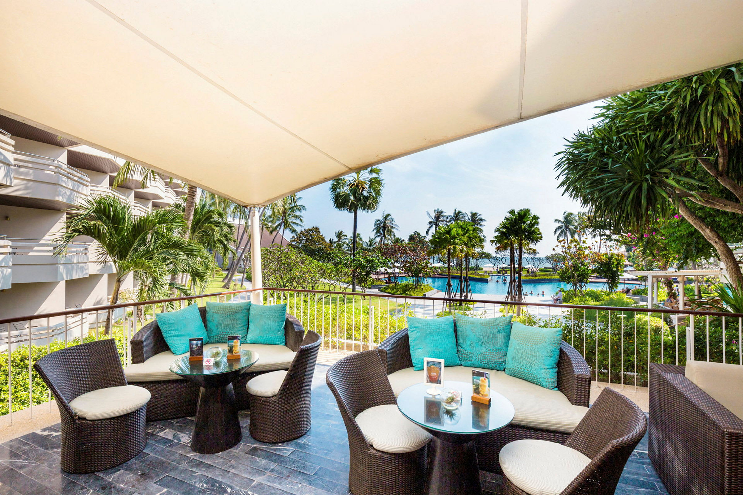 En overdækket terrasse med siddegrupper og borde af rattan samt udsigt over en pool og palmetræer. (AI-genereret tekst)