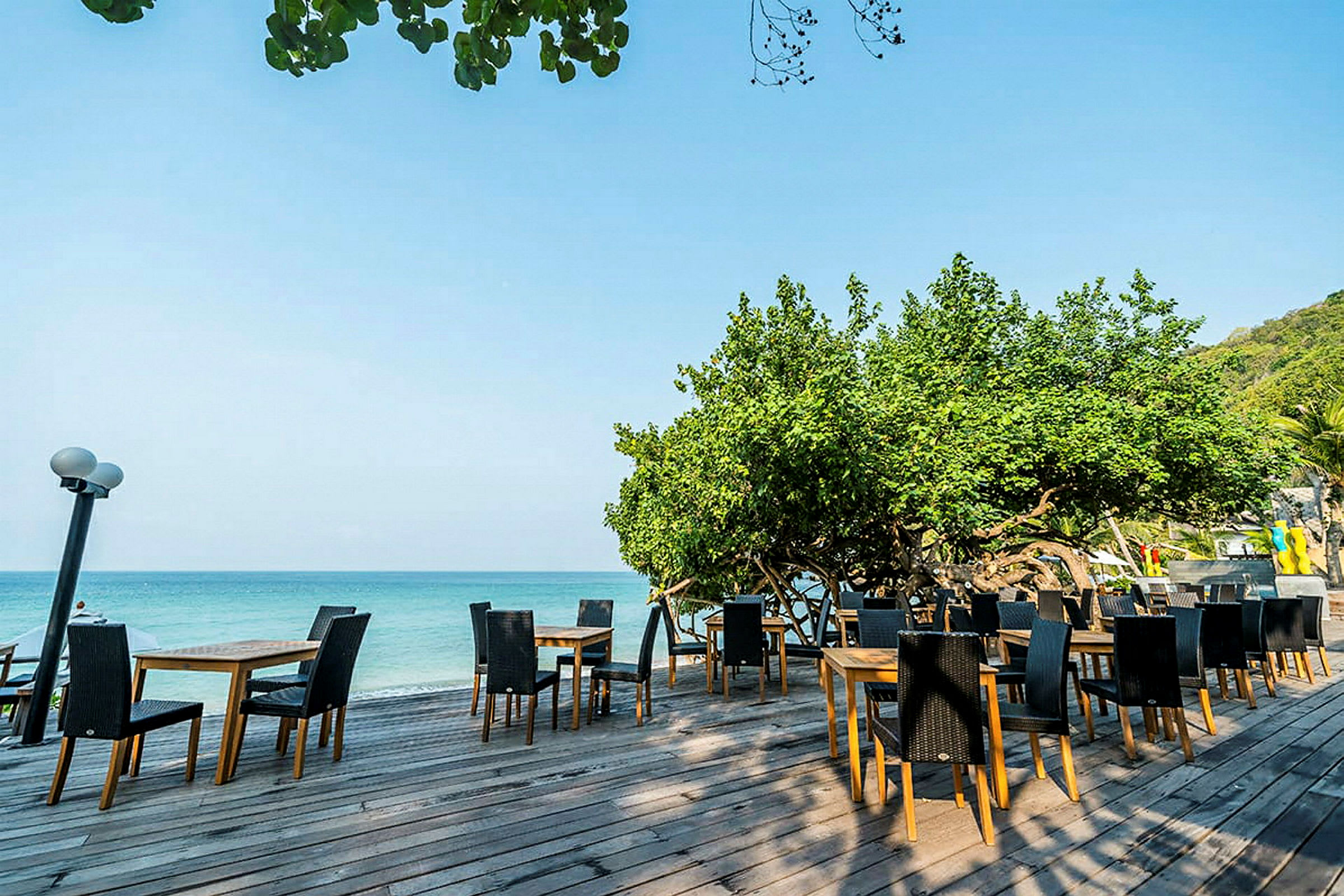 En udendørsrestaurant med borde og stole placeret på en træterrasse ved havet, omgivet af grønt. (AI-genereret tekst)