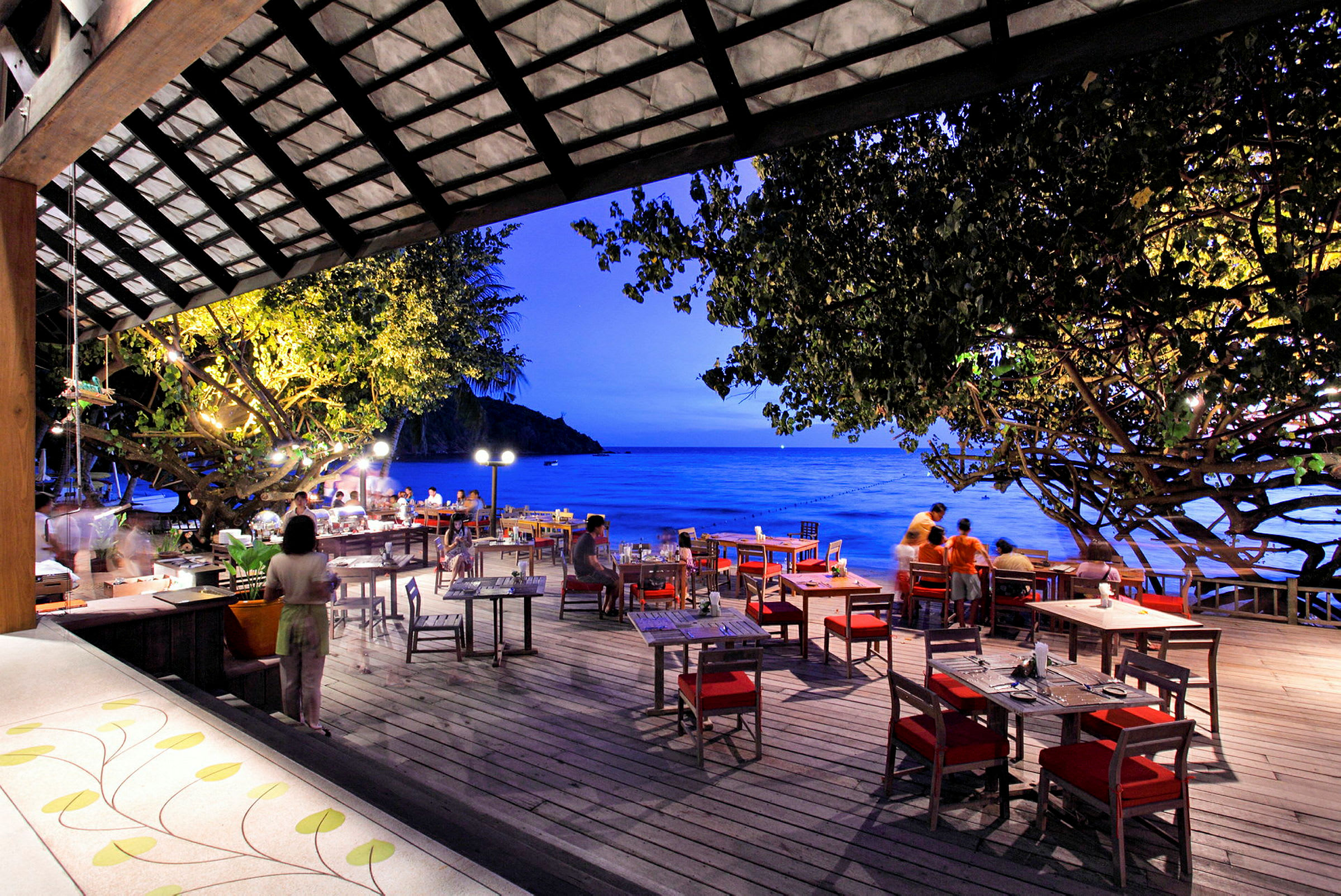 En restaurant med udendørs servering ved havet i aftenbelysning. Borde og stole er opstillet på en træterrasse. (AI-genereret tekst)
