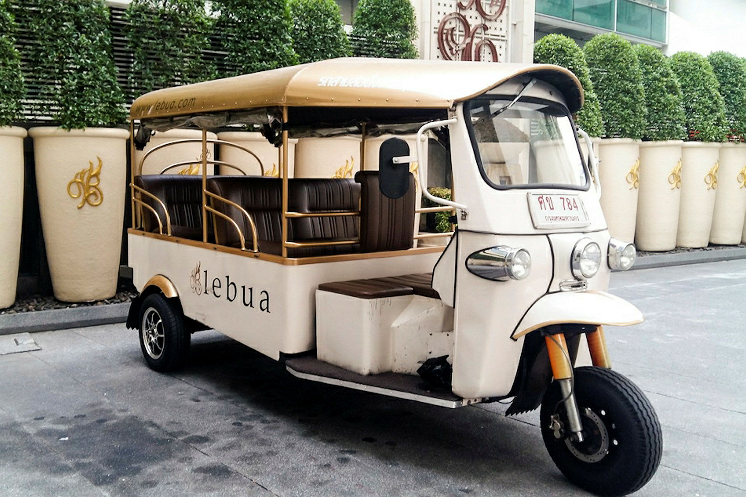En hvid tuk-tuk med beige tag står parkeret ved siden af en række store hvide krukker og grønne planter i baggrunden. (AI-genereret tekst)
