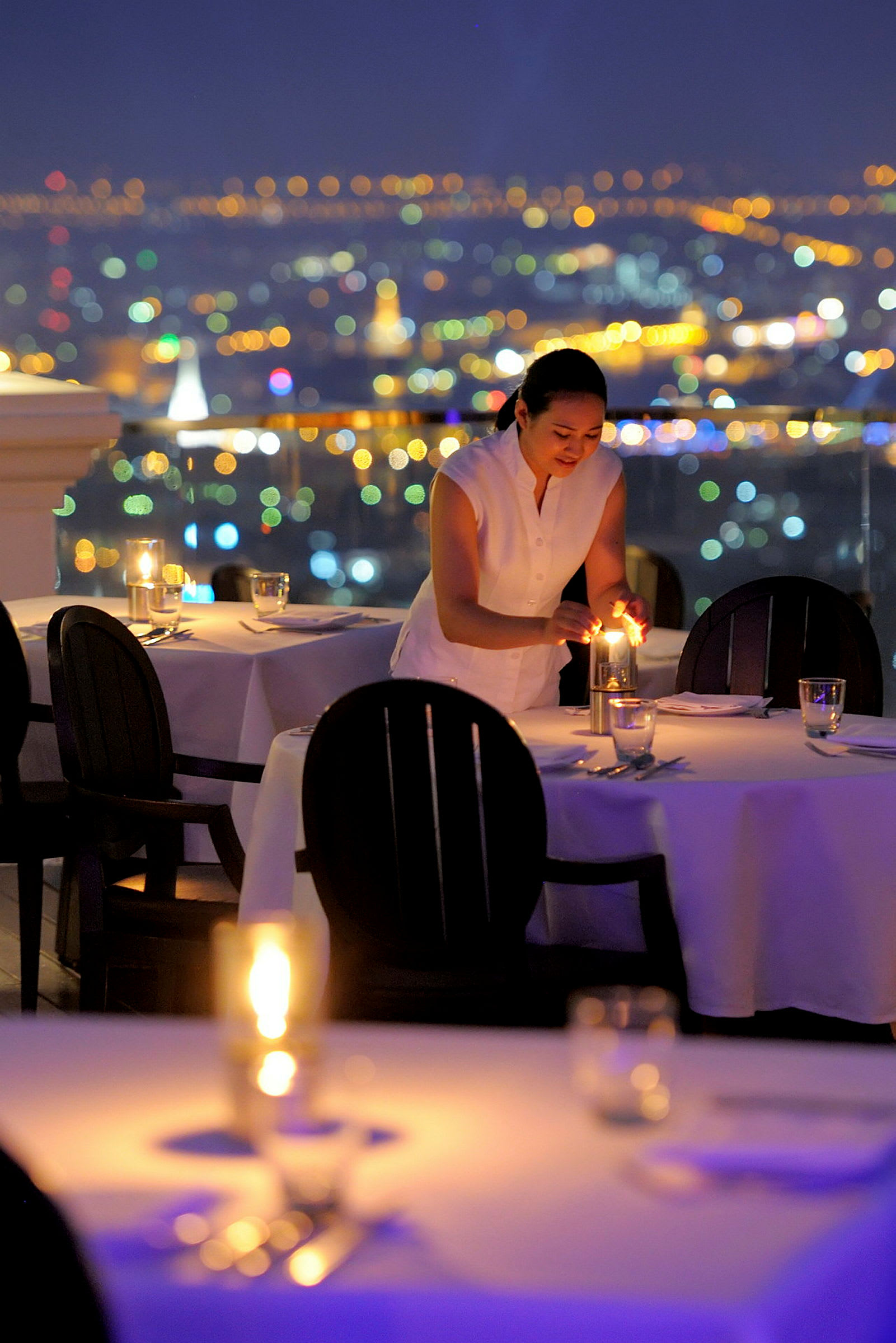 En person tænder lys på et dækket bord i en restaurant med udsigt over en bys skyline. (AI-genereret tekst)