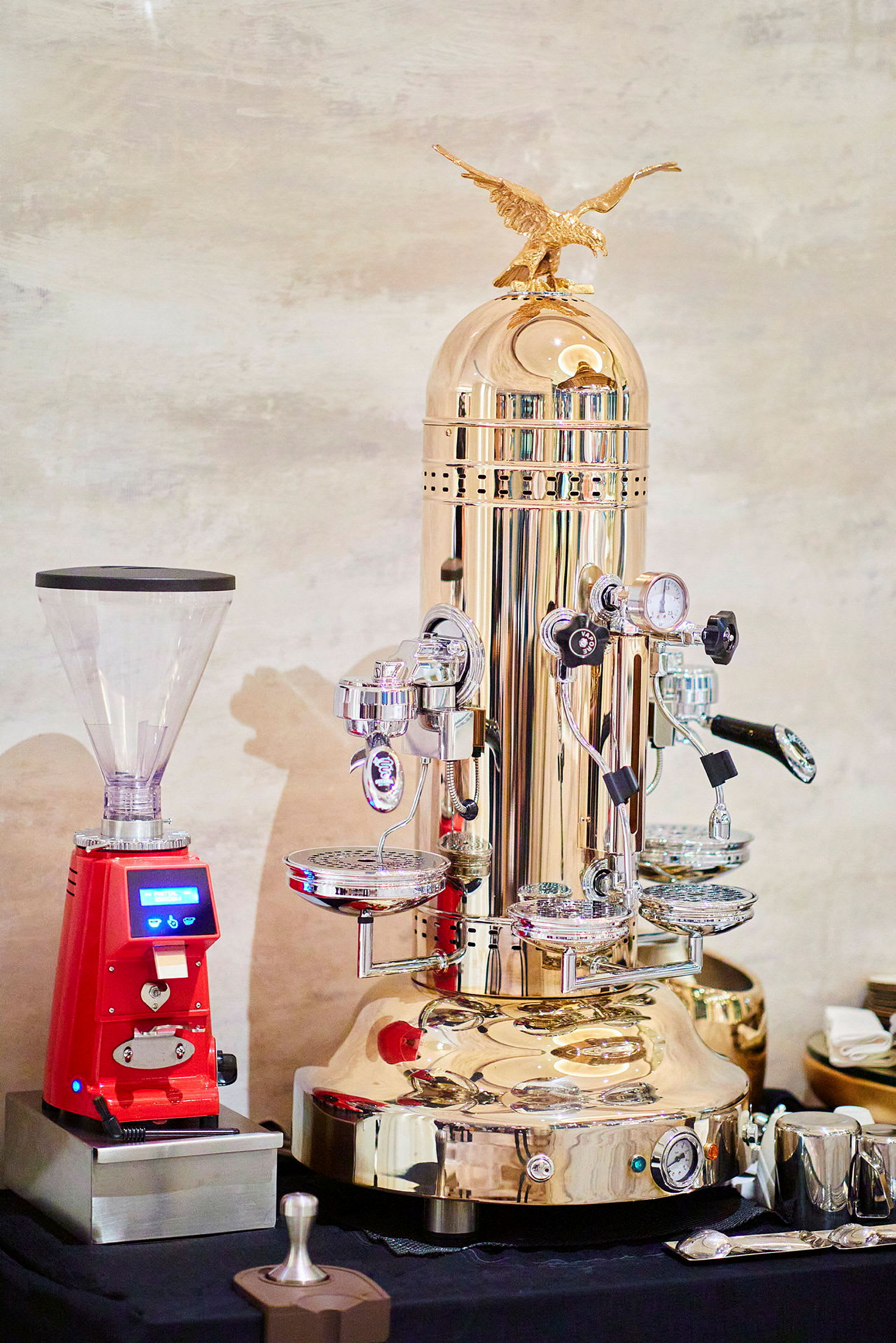 En elegant kaffemaskine i metallisk farve med flere bryggehoveder og detaljer ved siden af en rød kaffekværn. (AI-genereret tekst)