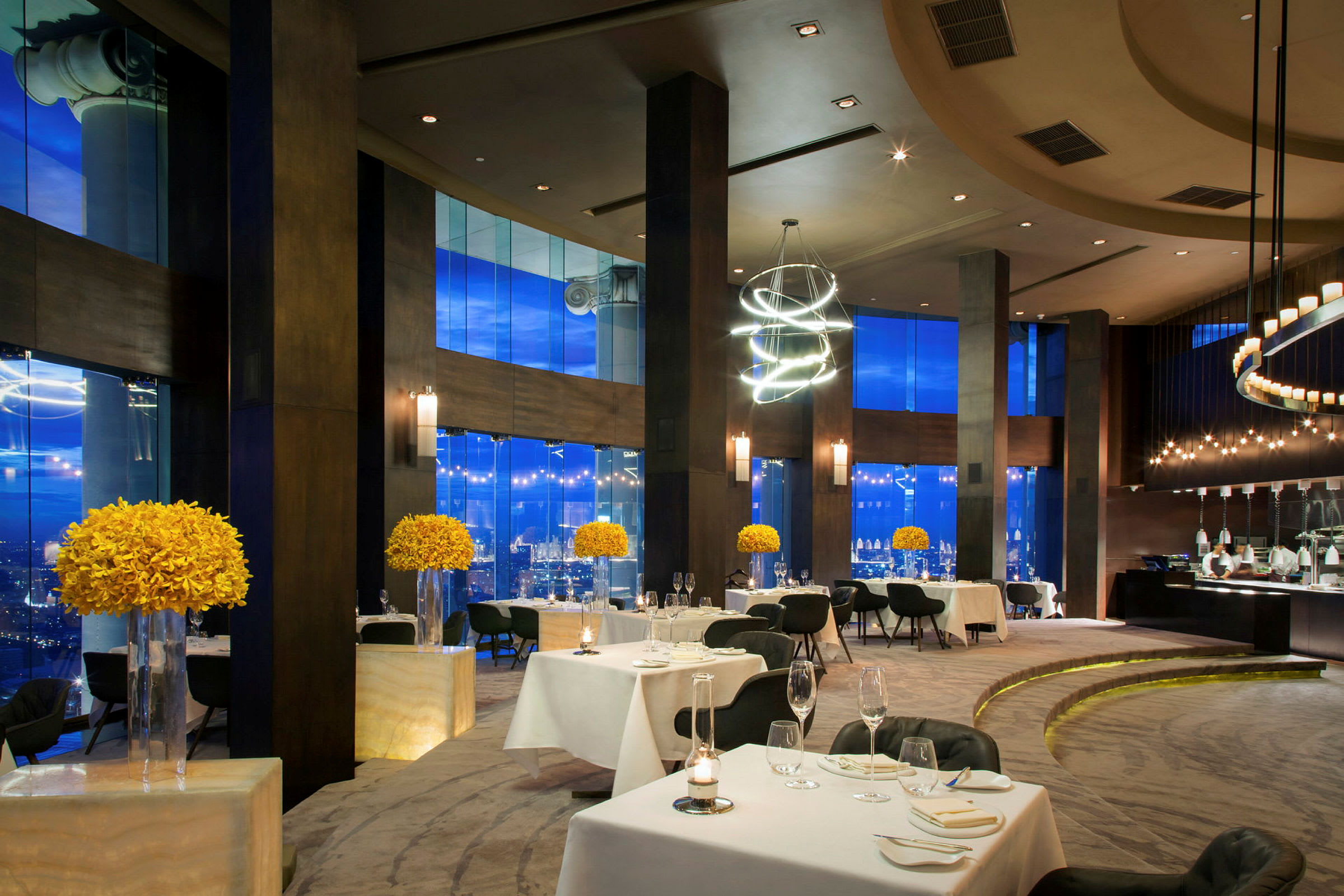 En elegant restaurant med dækkede borde, moderne møbler og store vinduer med udsigt til en by. Gule blomsterdekorationer er placeret på piedestaler. (AI-genereret tekst)