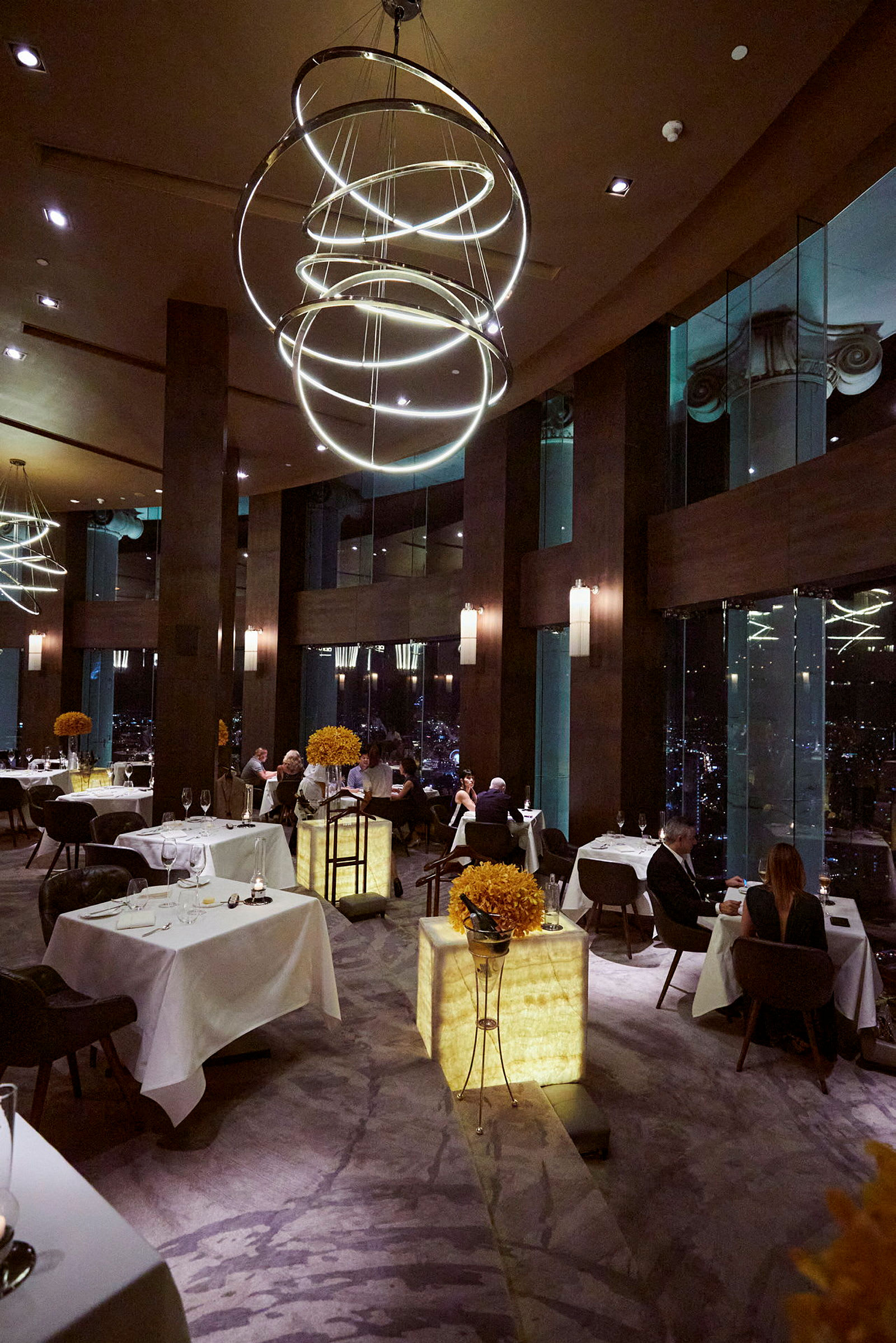 En luksuriøs restaurant med vinduer, der viser byudsigt om aftenen. I forgrunden ses borde med borddekorationer. (AI-genereret tekst)