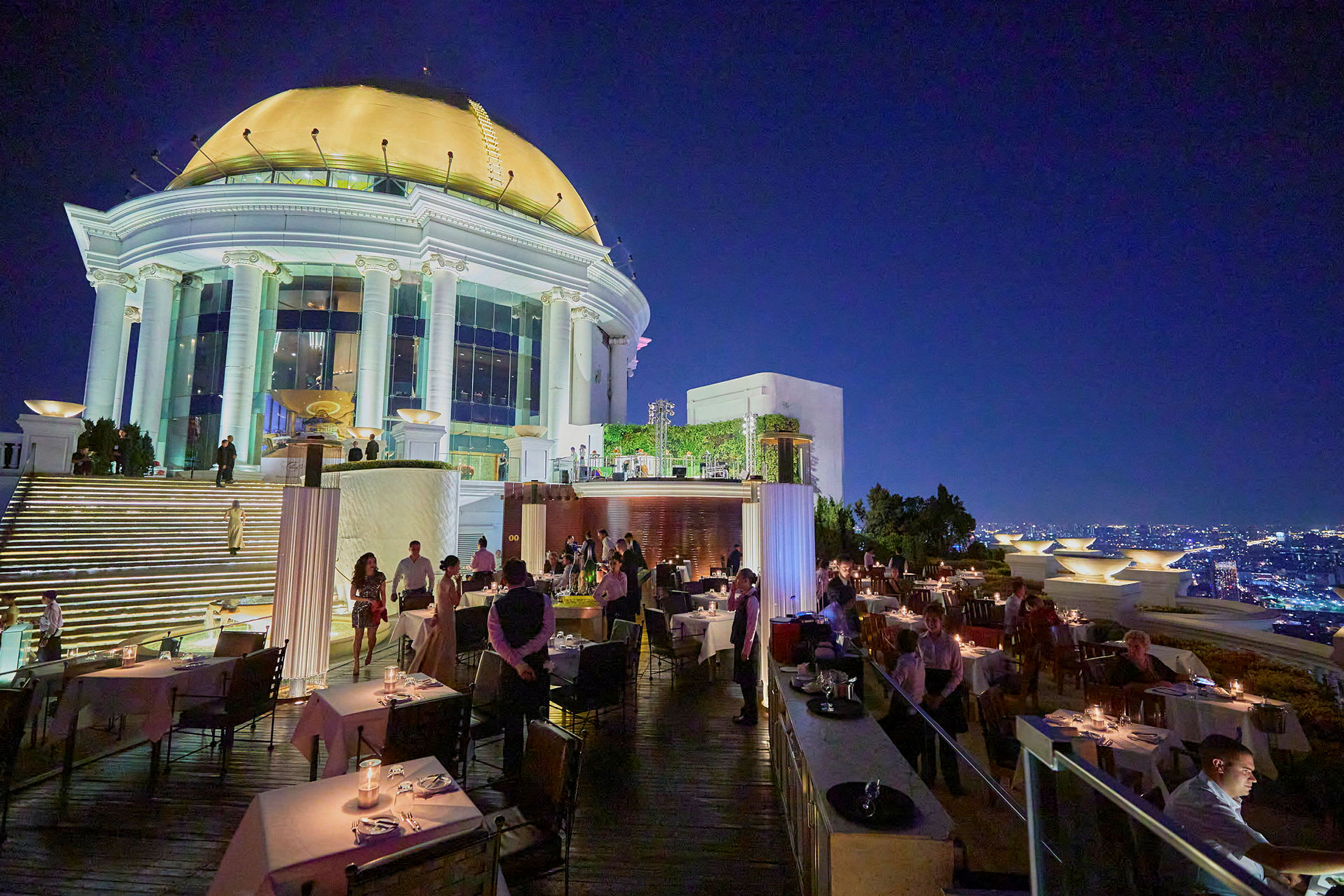 En restaurant på en tagterrasse med udsigt over en oplyst by i tusmørket. Billedet viser borde, gæster og tjenere. (AI-genereret tekst)