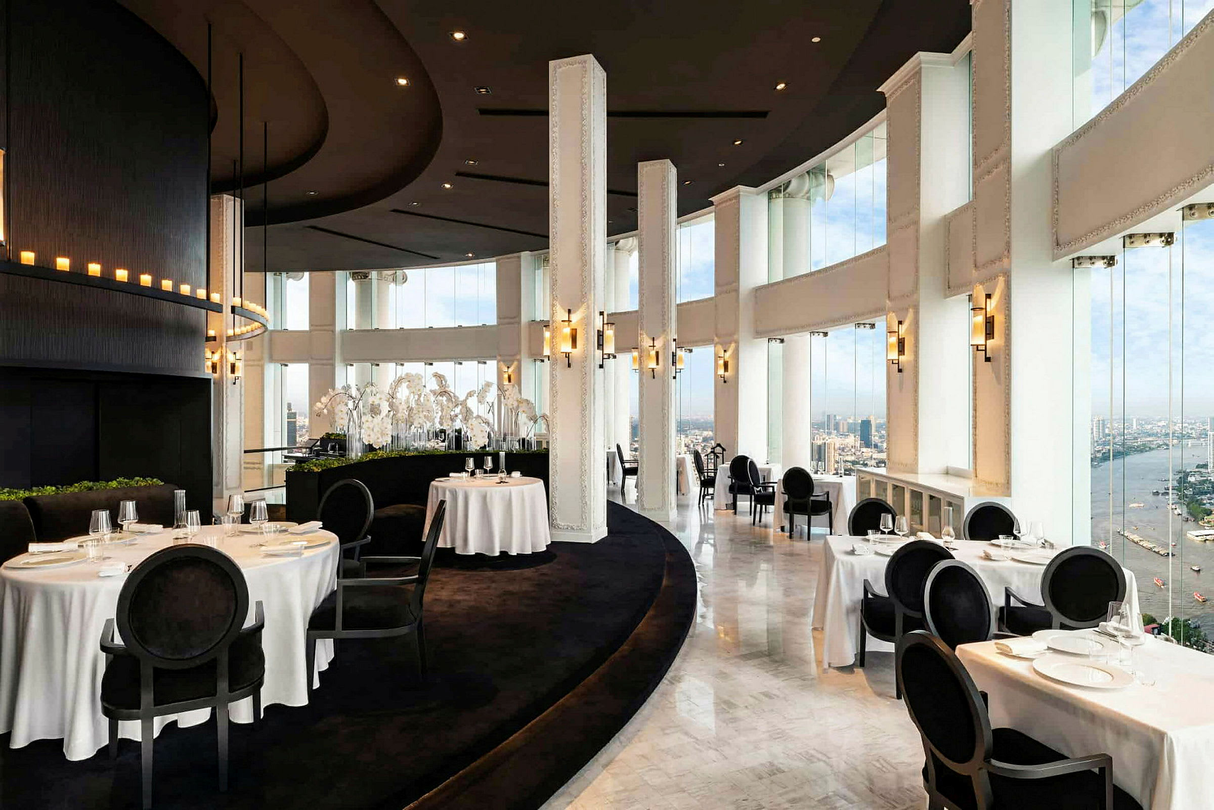 En elegant restaurant med store vinduer og hvid dugdækning, der giver udsigt over byen og en flod. (AI-genereret tekst)