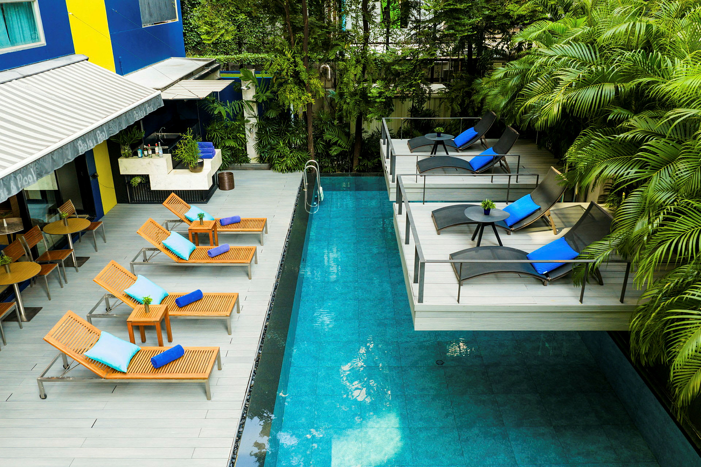 En swimmingpool med tilhørende terrasse og liggestole udstyret med puder og håndklæder. Der er også omkringliggende vegetation. (AI-genereret tekst)