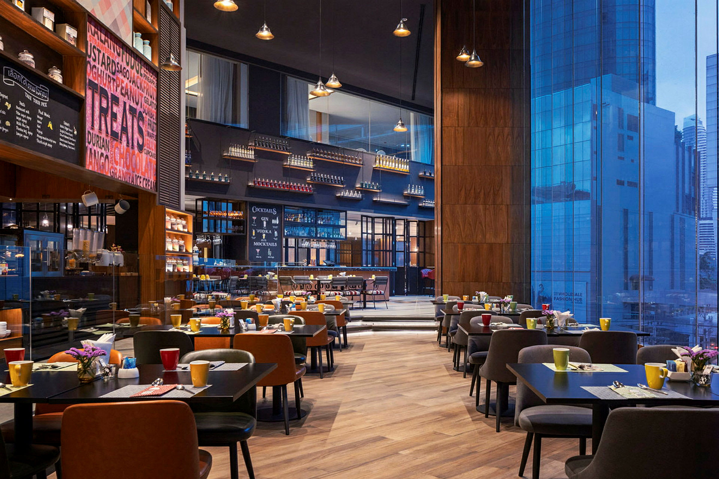 En rummelig restaurant med moderne borde og stole opstillet, farverige kopper og blomster på bordene samt en stor glasvæg med udsigt til byens bygninger. (AI-genereret tekst)