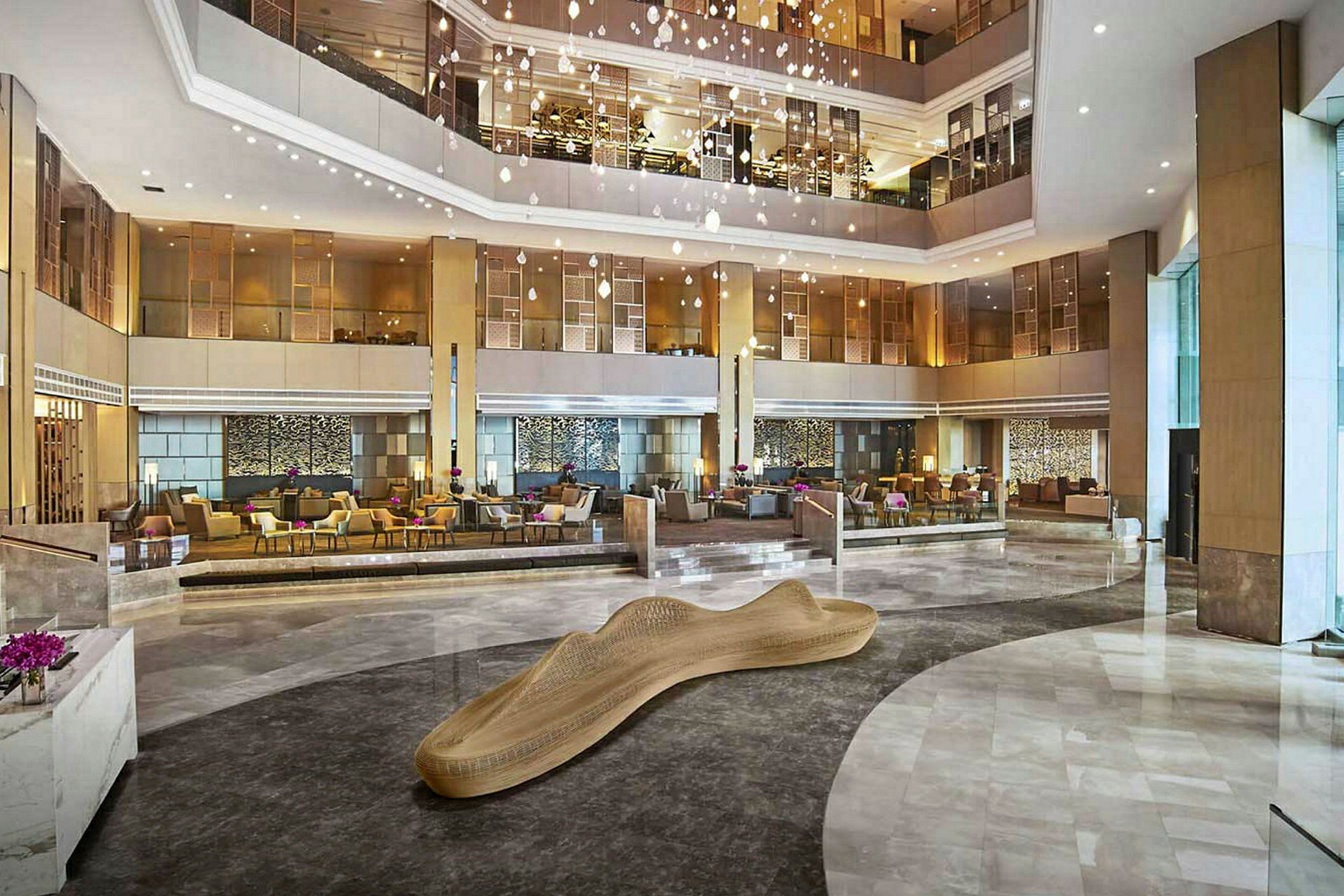 Et stort hotellobbyområde med moderne møbler og en dekorativ træskulptur i midten. Flere siddegrupper og elegant belysning. (AI-genereret tekst)
