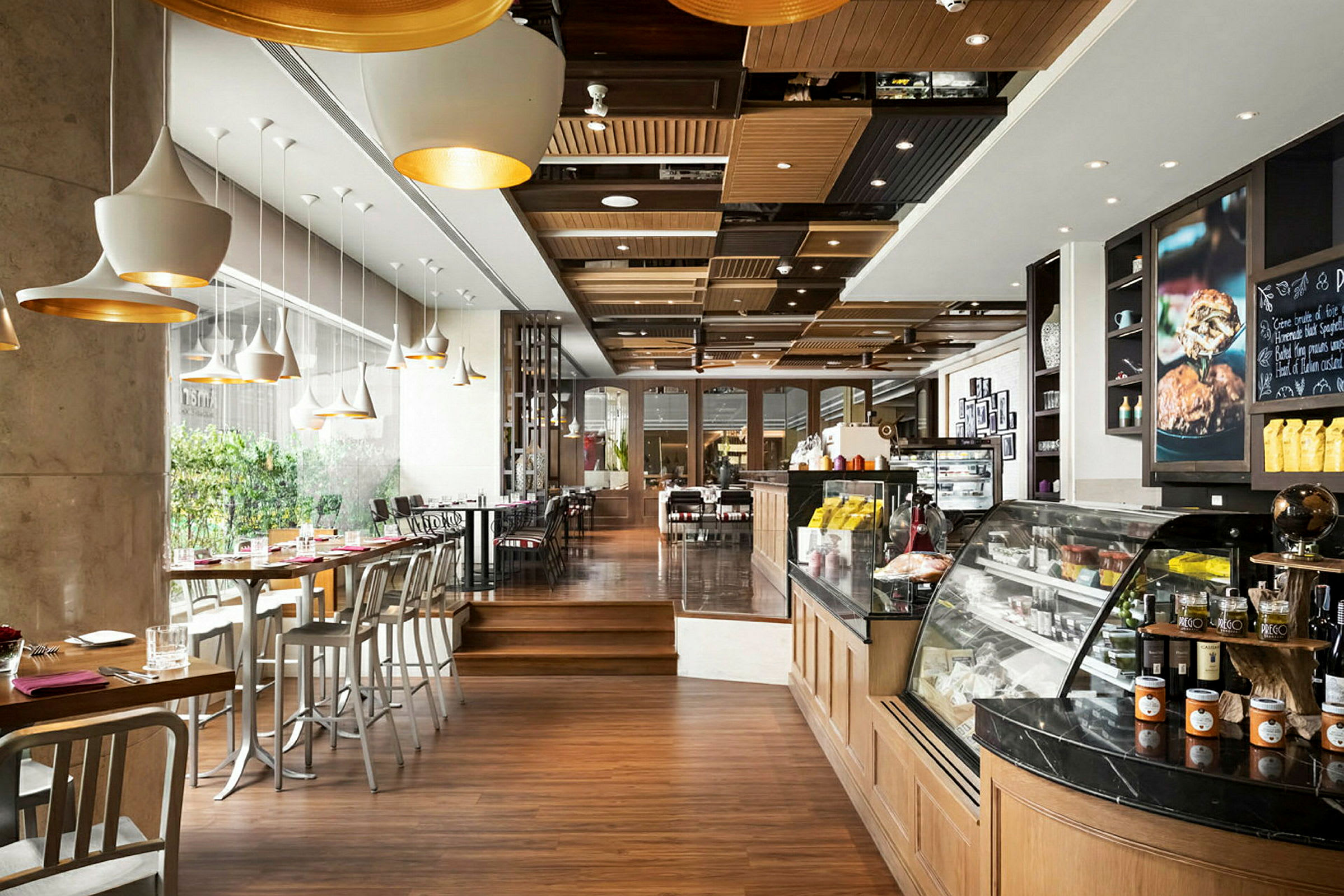 En moderne café med trægulv, store vinduer og siddepladser ved borde og barstole. En oplyst disk med mad og drikke findes til højre. (AI-genereret tekst)