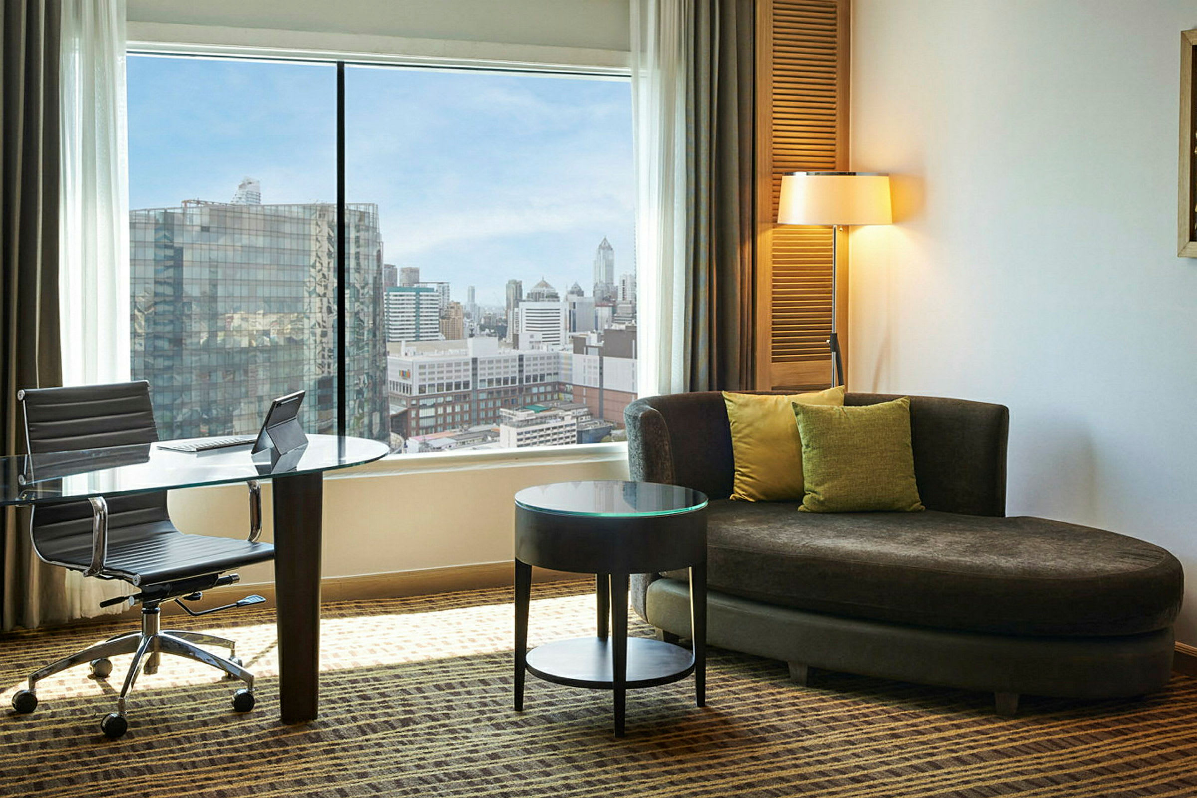Et hotelværelse med stor vinduesudsigt over en by. Der er et glasbord og stol ved vinduet samt en brun sofa med puder og et rundt sidebord. (AI-genereret tekst)