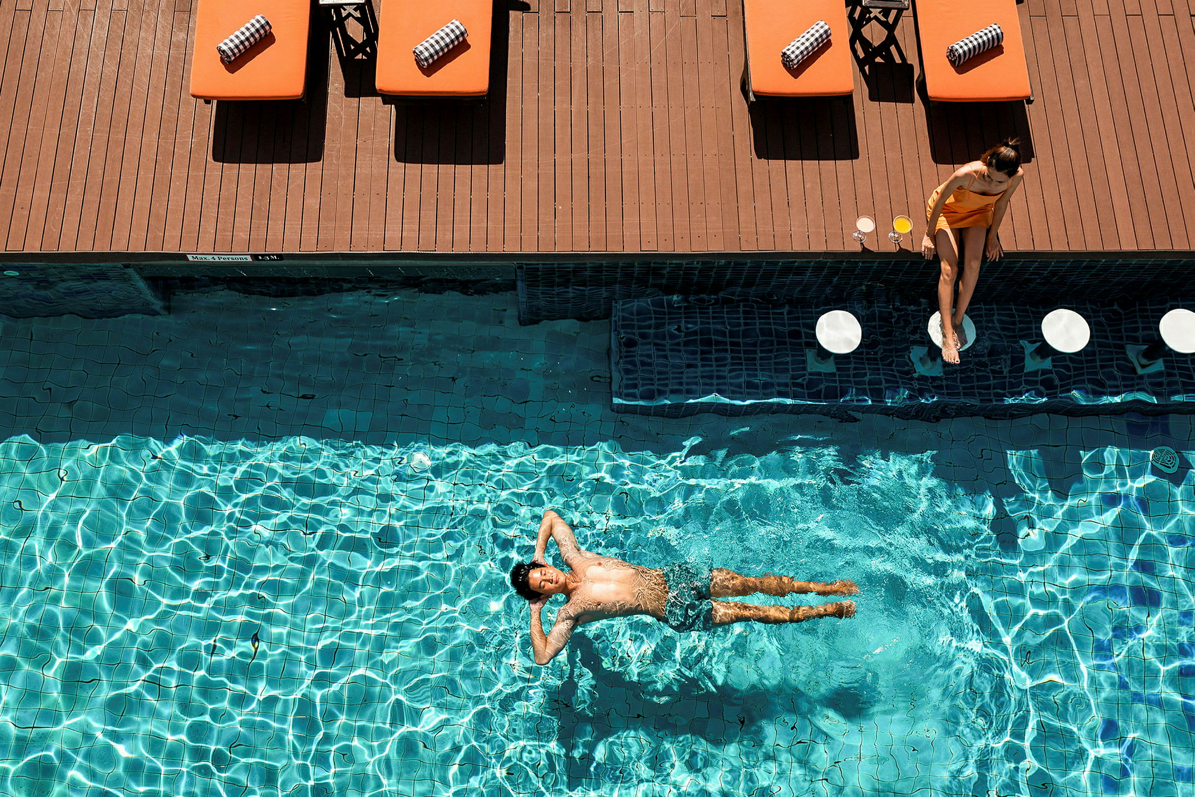 En person ligger og flyder i en pool, mens liggestole står langs kanten. (AI-genereret tekst)