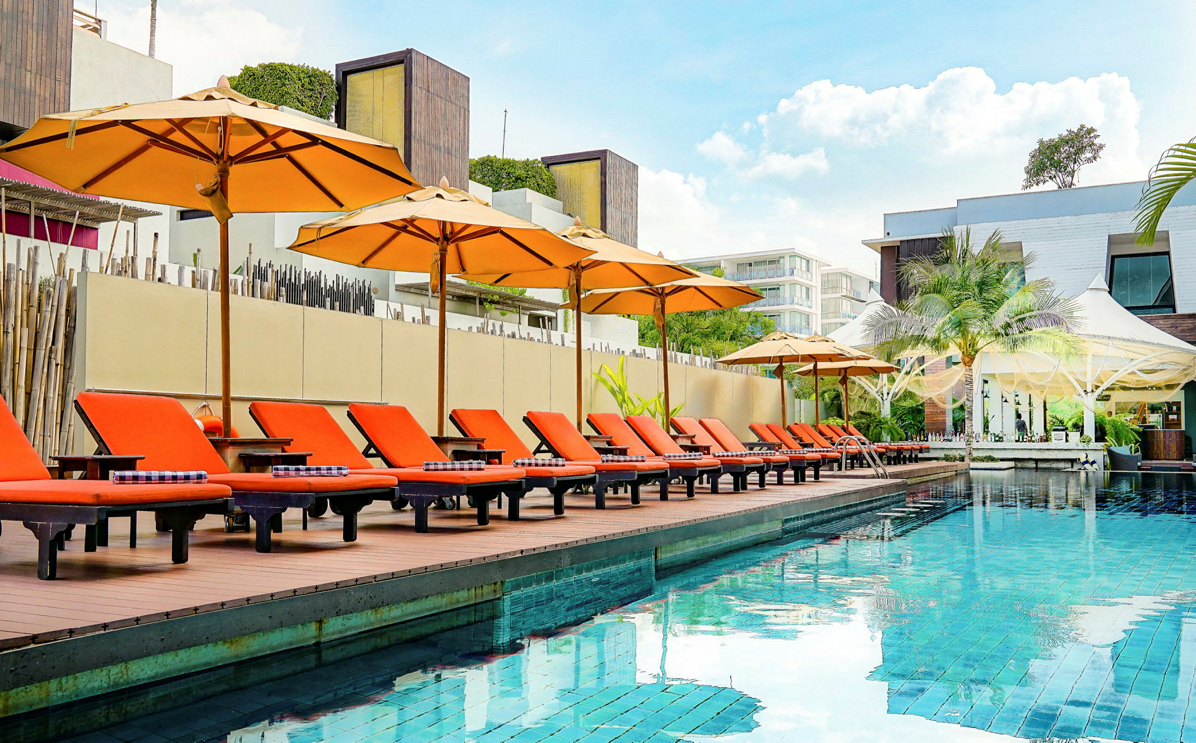 En udendørs pool med orange solsenge og parasoller ved kanten, omgivet af moderne arkitektur. (AI-genereret tekst)