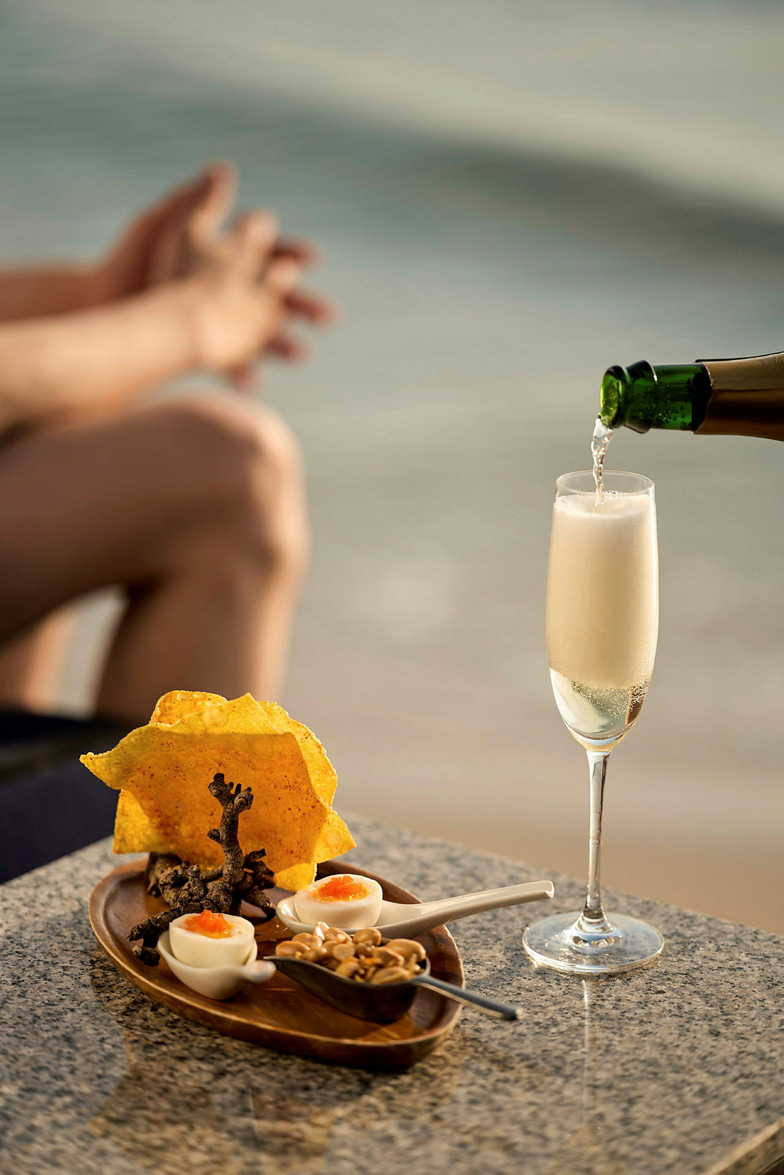 Et glas champagne bliver hældt op på et bord med en lille servering af snacks foran en sløret baggrund af strand og hav. (AI-genereret tekst)