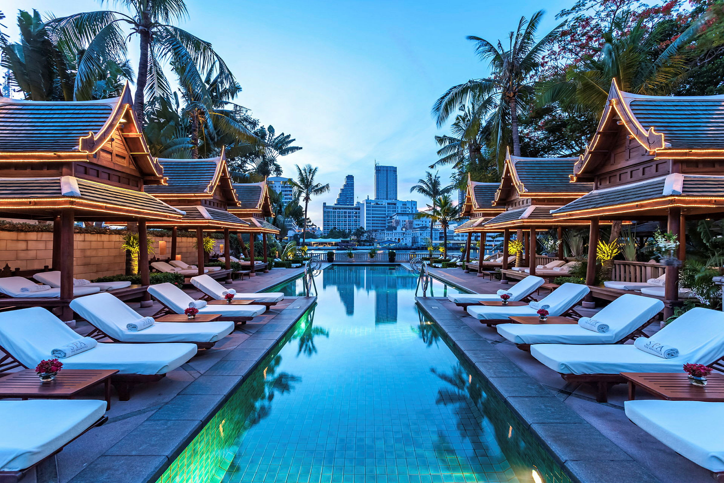 En pool med liggestole på begge sider, omgivet af pavilloner med thailandsk arkitektur og palmer, aftenbelysning. (AI-genereret tekst)