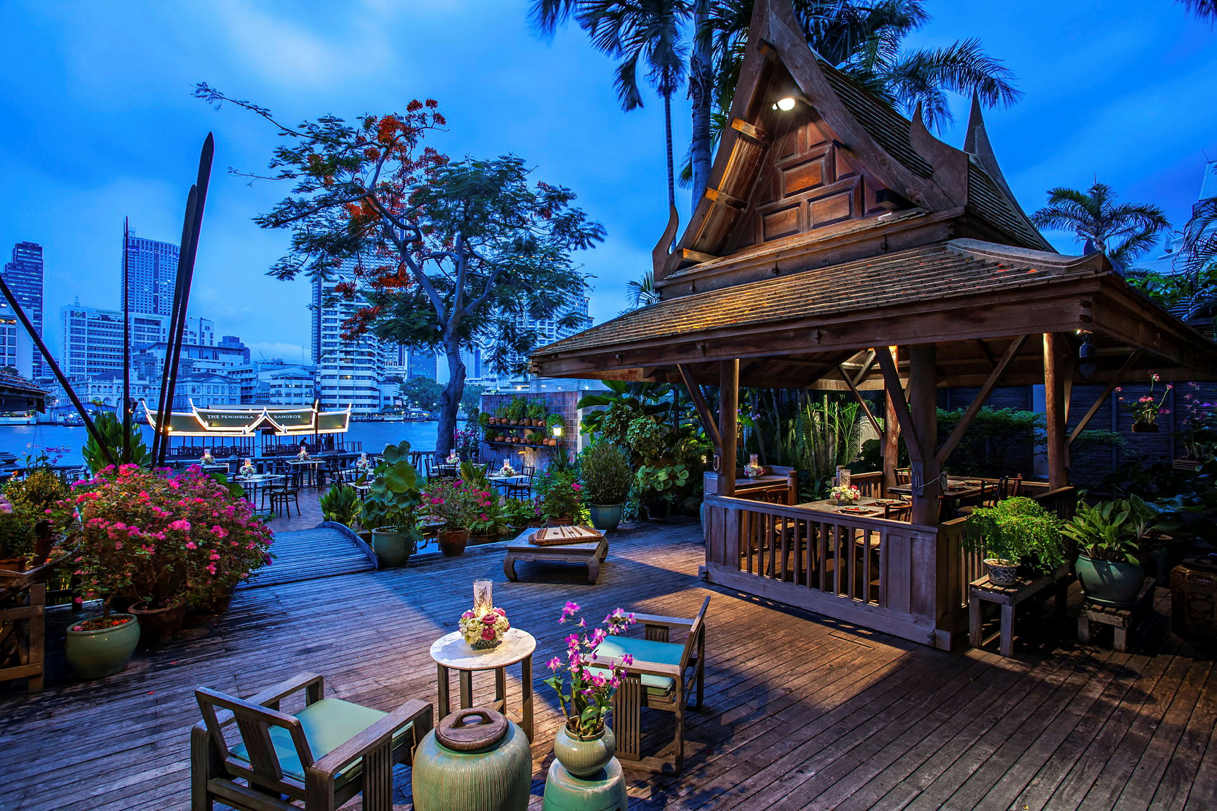 Et oplyst træhus med traditionelt thailandsk design står på en terrasse omgivet af blomster og planter. I baggrunden ses en flod og byens skyline ved skumringen. (AI-genereret tekst)
