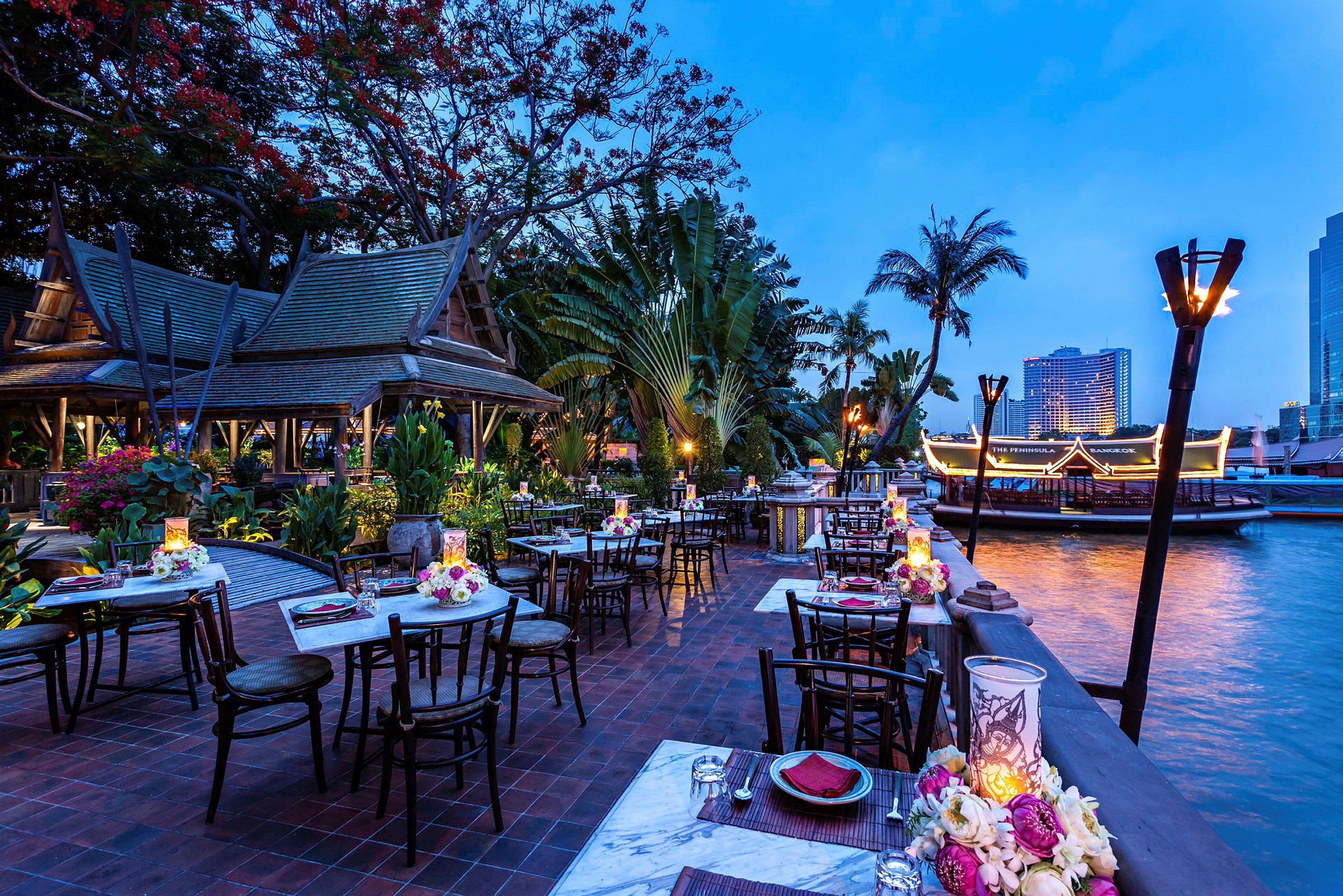 En udendørsrestaurant med opdækkede borde ved en flod. Restauranten har traditionelle thailandske detaljer og er omgivet af frodig vegetation. (AI-genereret tekst)