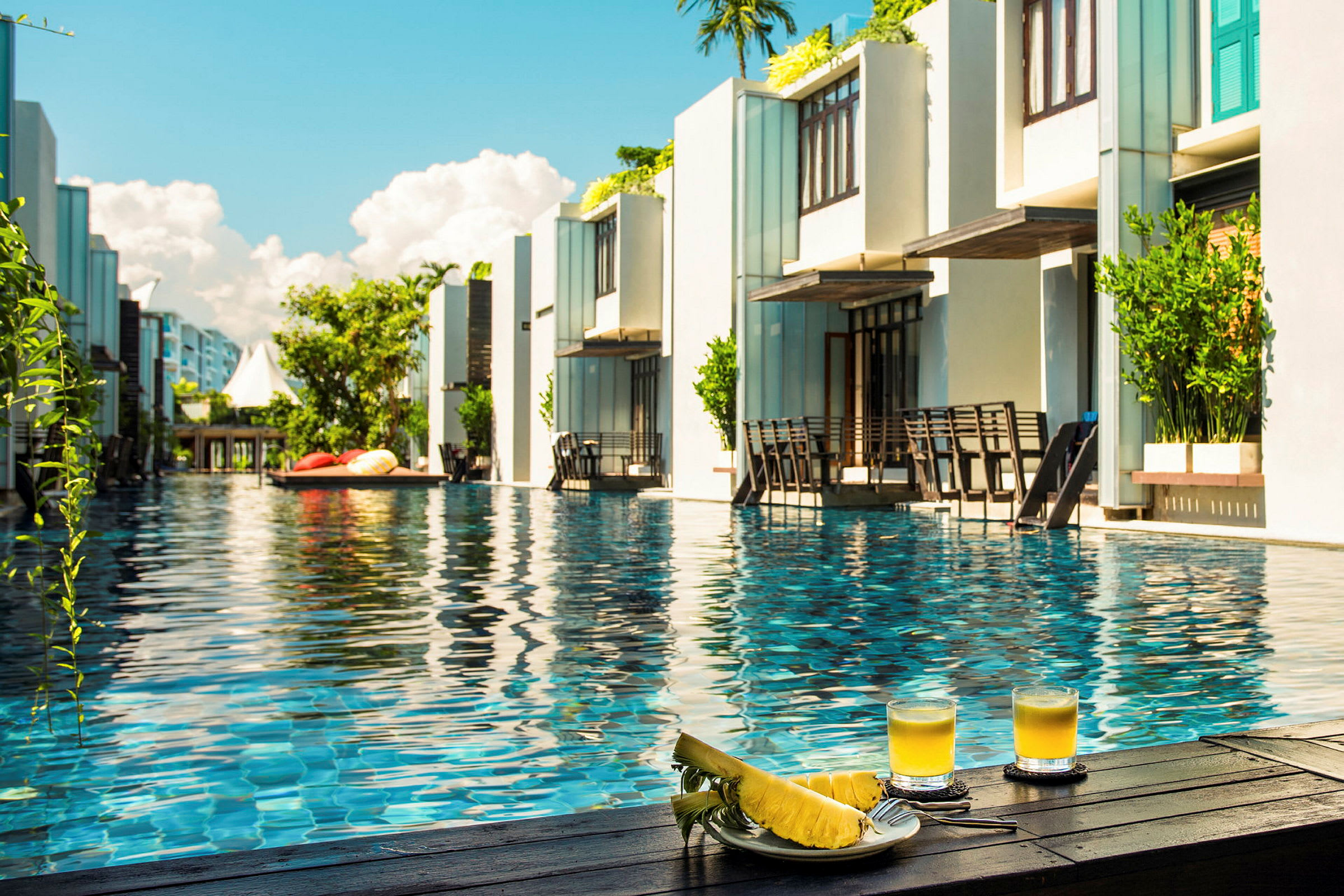 Resortpool omgivet af moderne bygninger med to glas juice og skåret ananas i forgrunden. (AI-genereret tekst)
