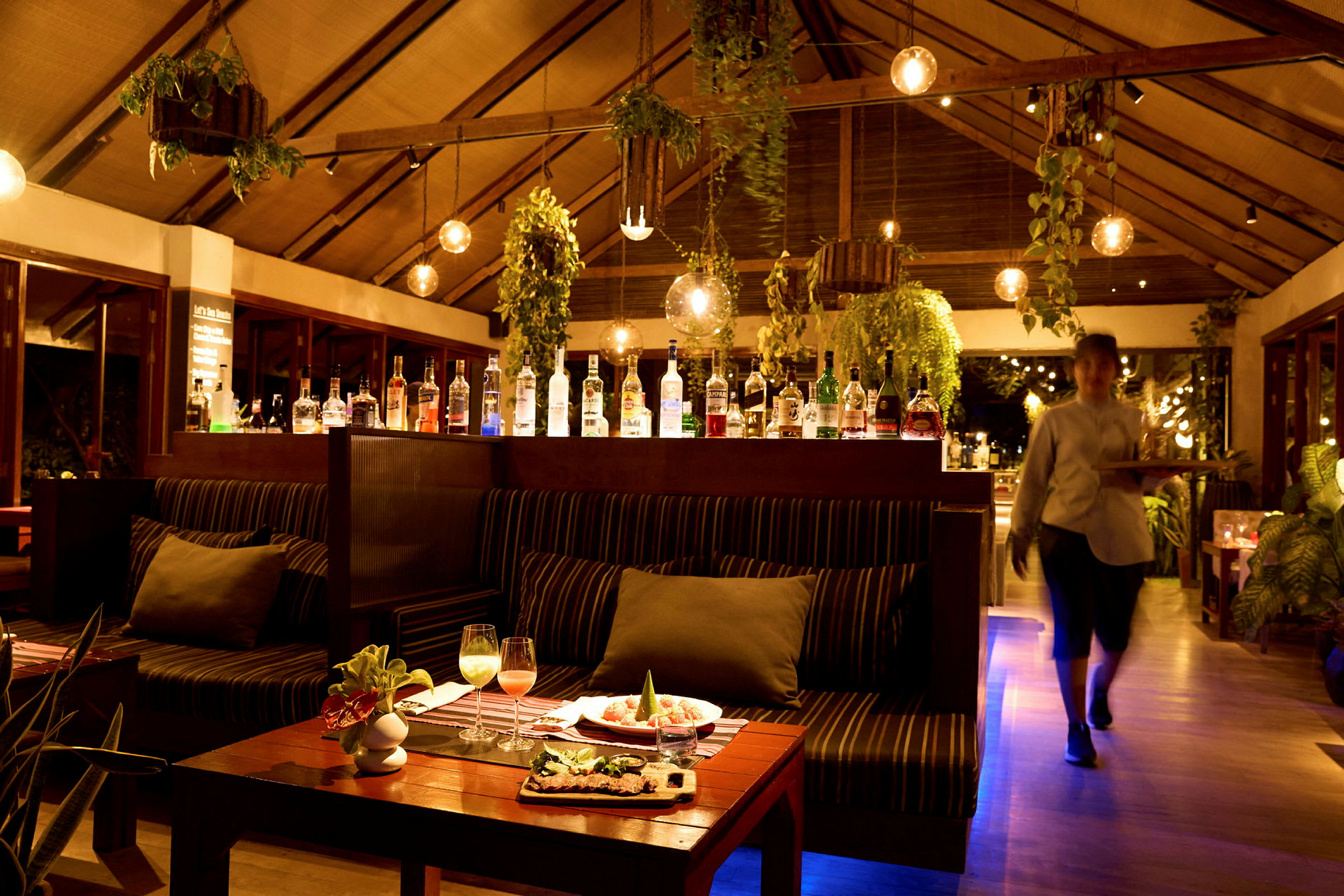 Interiør af en restaurant med siddepladser, hængende planter og en bar med forskellige spiritusflasker. (AI-genereret tekst)
