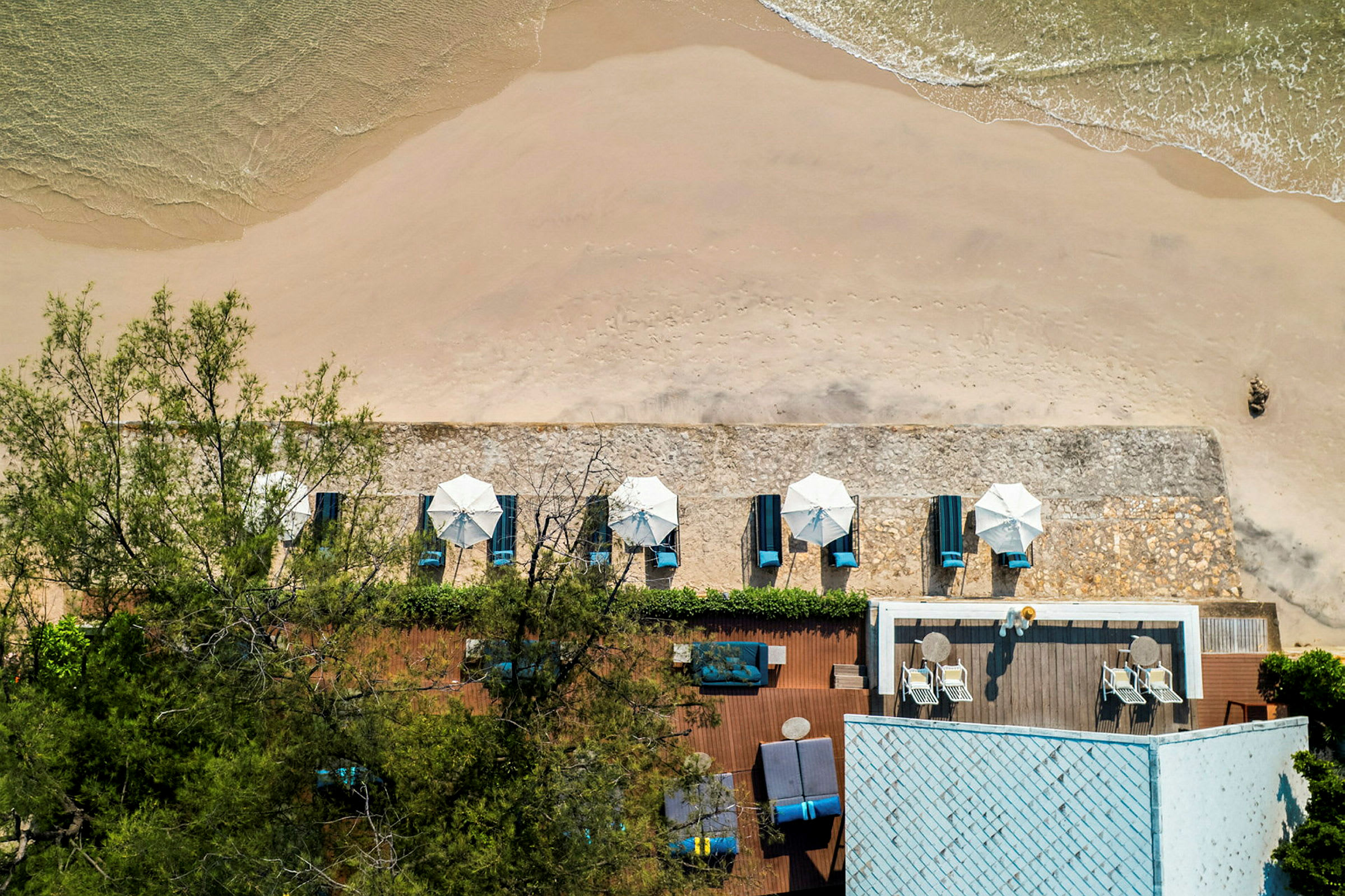 Udsigt ovenfra af en sandstrand med parasoller og liggestole samt en nærliggende bygning med terrasse. (AI-genereret tekst)