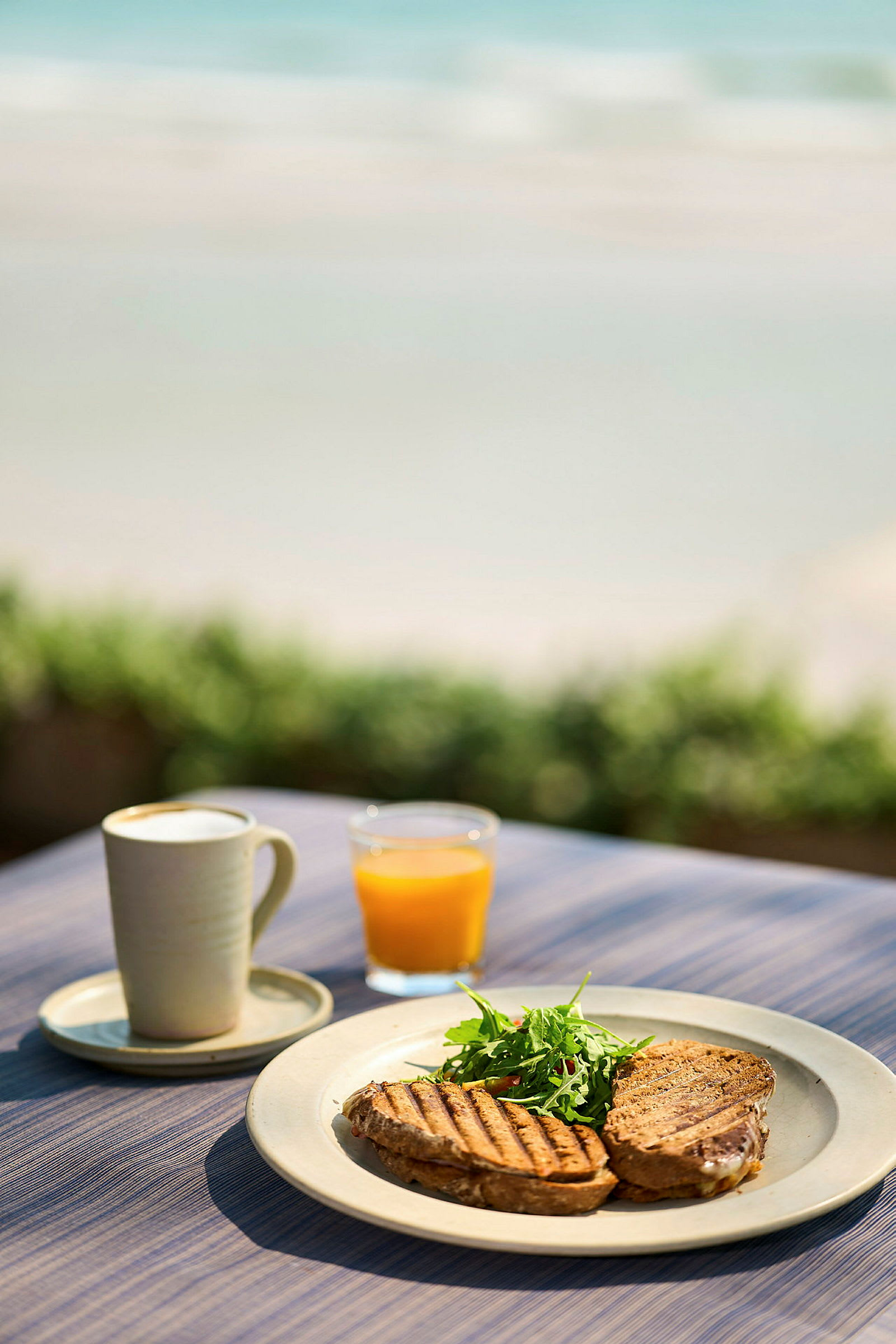 En tallerken med grillede sandwiches og salat, et glas appelsinjuice og en kop kaffe står på et bord ved stranden. (AI-genereret tekst)