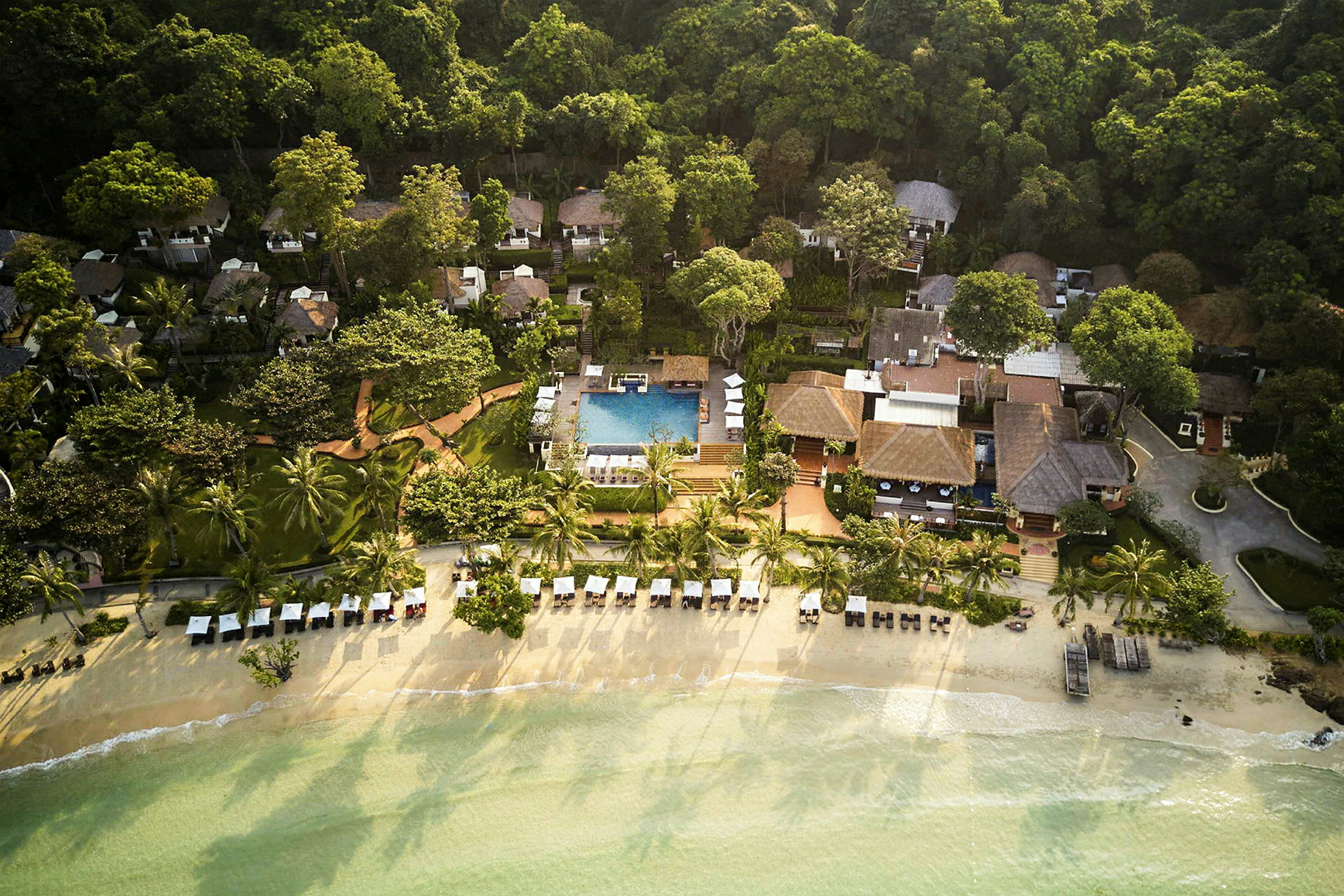 Luftfoto af en ferieresort ved stranden med palmer, liggestole og en pool, omgivet af frodigt grønt. (AI-genereret tekst)