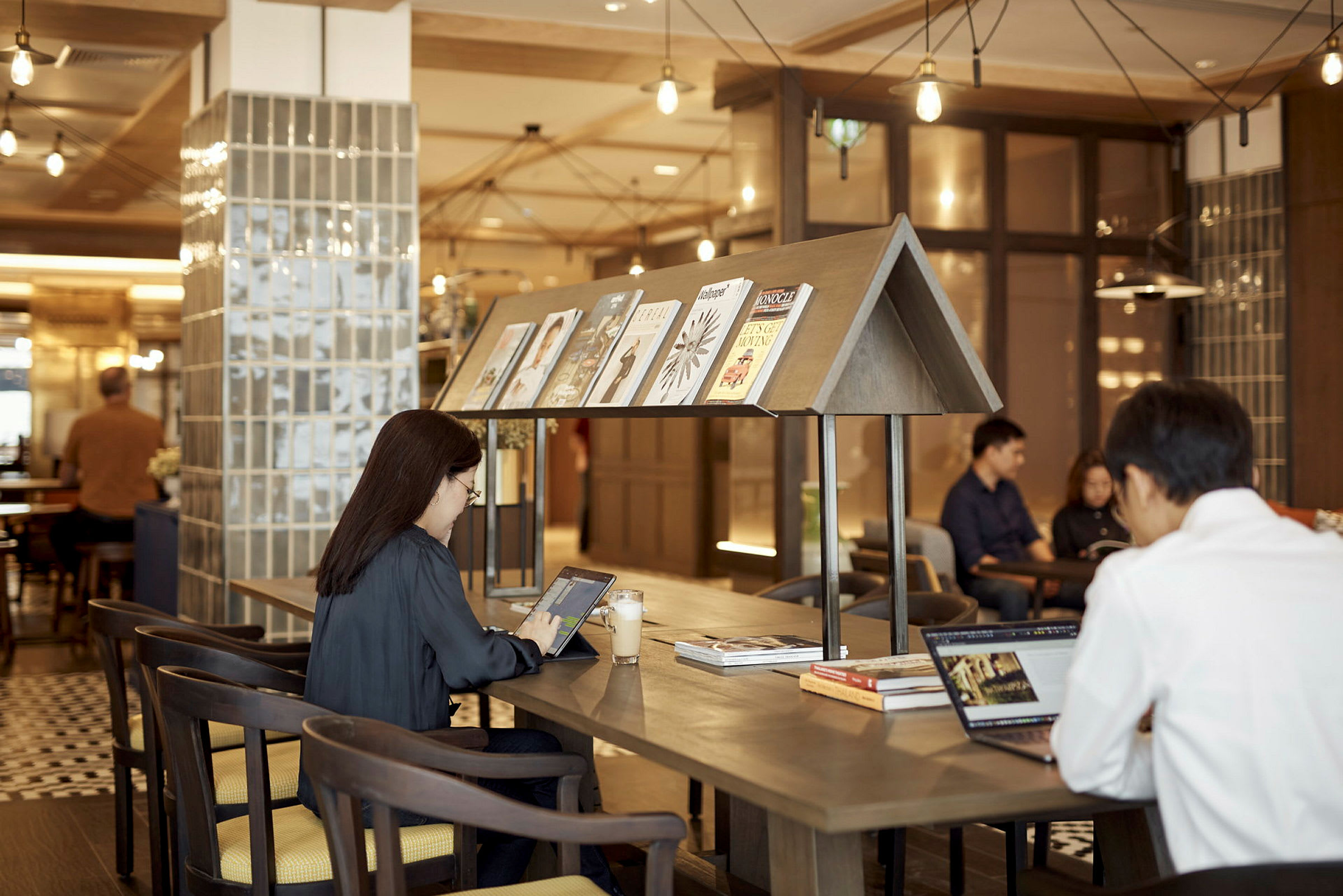 Et moderne café- eller arbejdsområde med siddepladser og borde. På bordet ses tidsskrifter og personer, der arbejder på bærbare computere. (AI-genereret tekst)