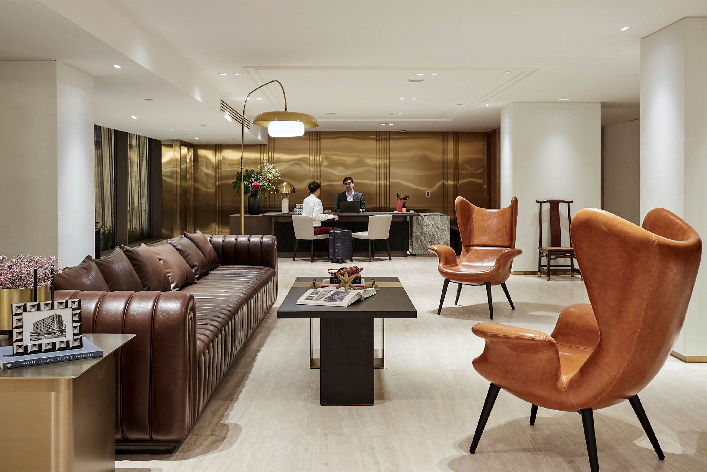Et hotellobbyområde med en brun lædersofa, orange læderlænestole og et mørkt sofabord foran. I baggrunden er en receptionsdisk, hvor to personer taler sammen. (AI-genereret tekst)