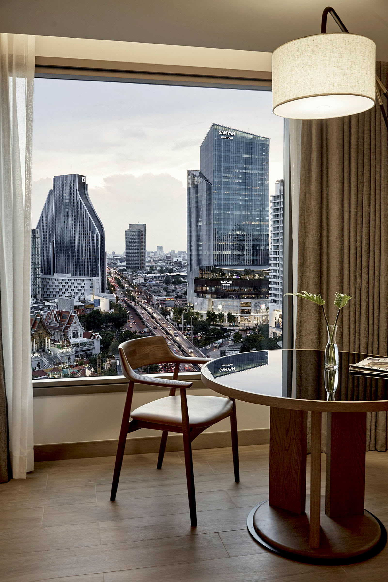 Et moderne hotelværelse med udsigt over en by med høje bygninger i aftenlys. Et vindue viser udsigten, og foran det står et træbord med en stol og en enkel blomsterpotte. (AI-genereret tekst)