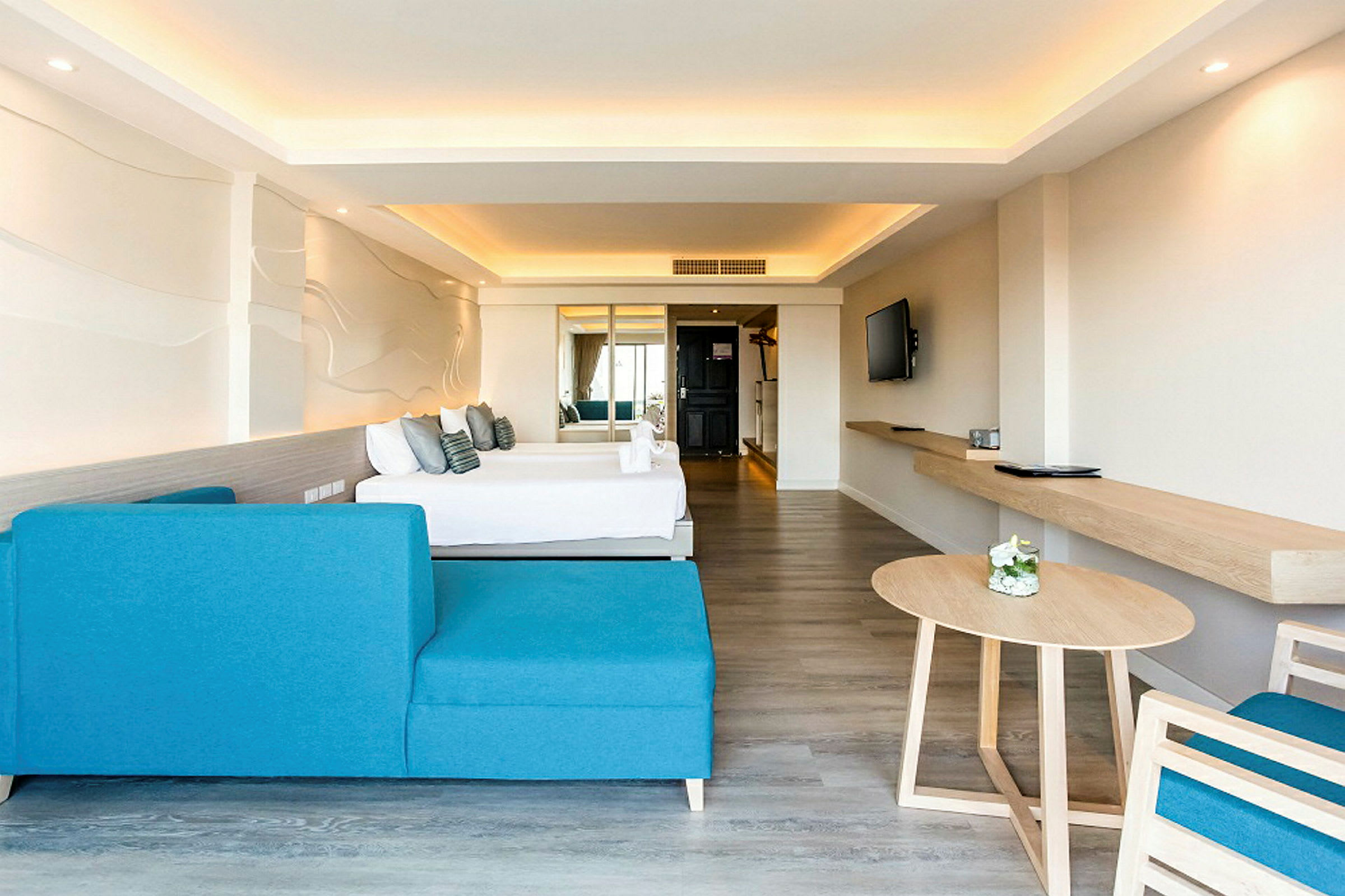 Et moderne hotelværelse med en blå sofa, et bord og en seng i baggrunden. Væggen har et dekorativt design, og rummet er lyst med neutrale farver. (AI-genereret tekst)