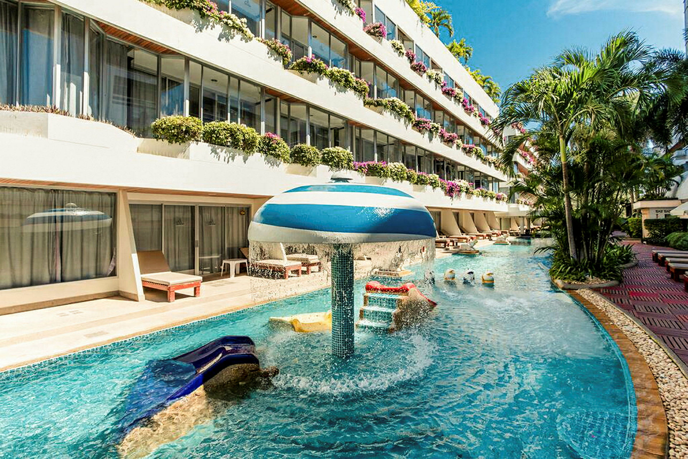 En pool med vandattraktion ved et hotel med flere etager og altaner fyldt med blomster. (AI-genereret tekst)