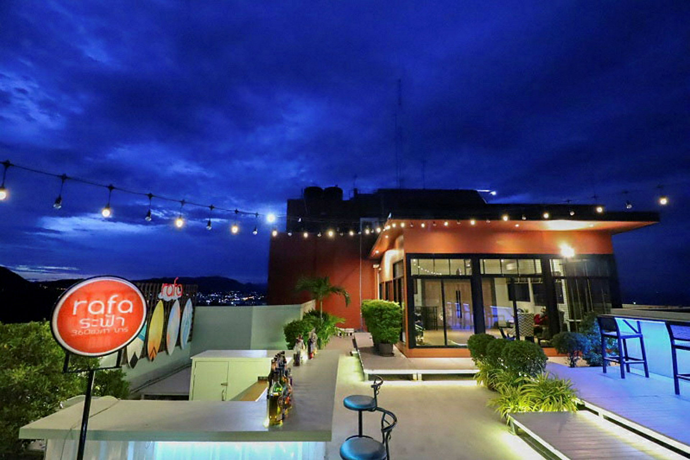 En tagterrasse med en bar og stole. Lys hænger over, og i baggrunden ses en bygning og en by med nattehimmel. (AI-genereret tekst)