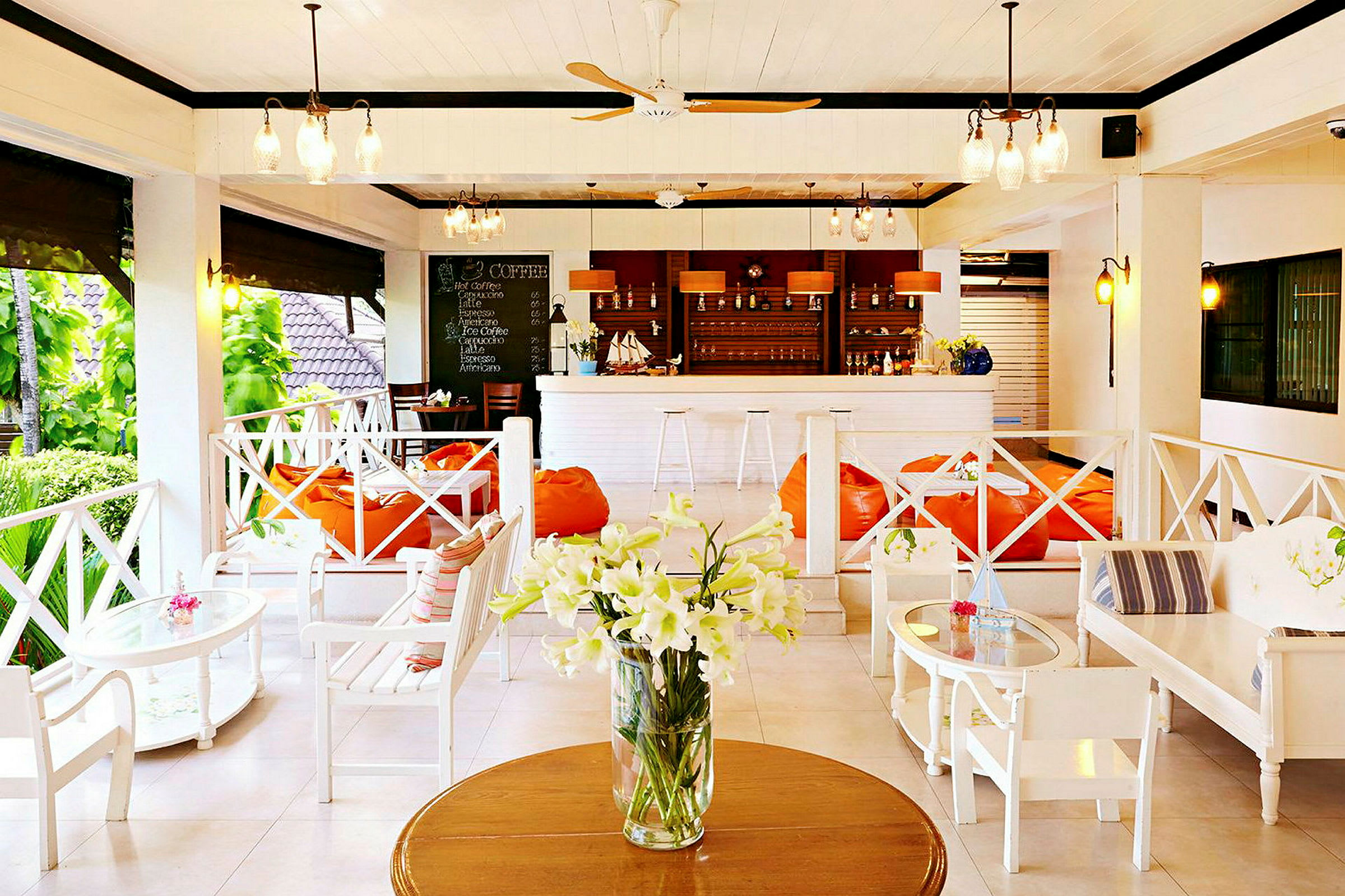 En lys og åben lounge med hvide møbler, orange sækkestole og en bar med drikkevarer i baggrunden samt en vase med hvide blomster på et bord. (AI-genereret tekst)