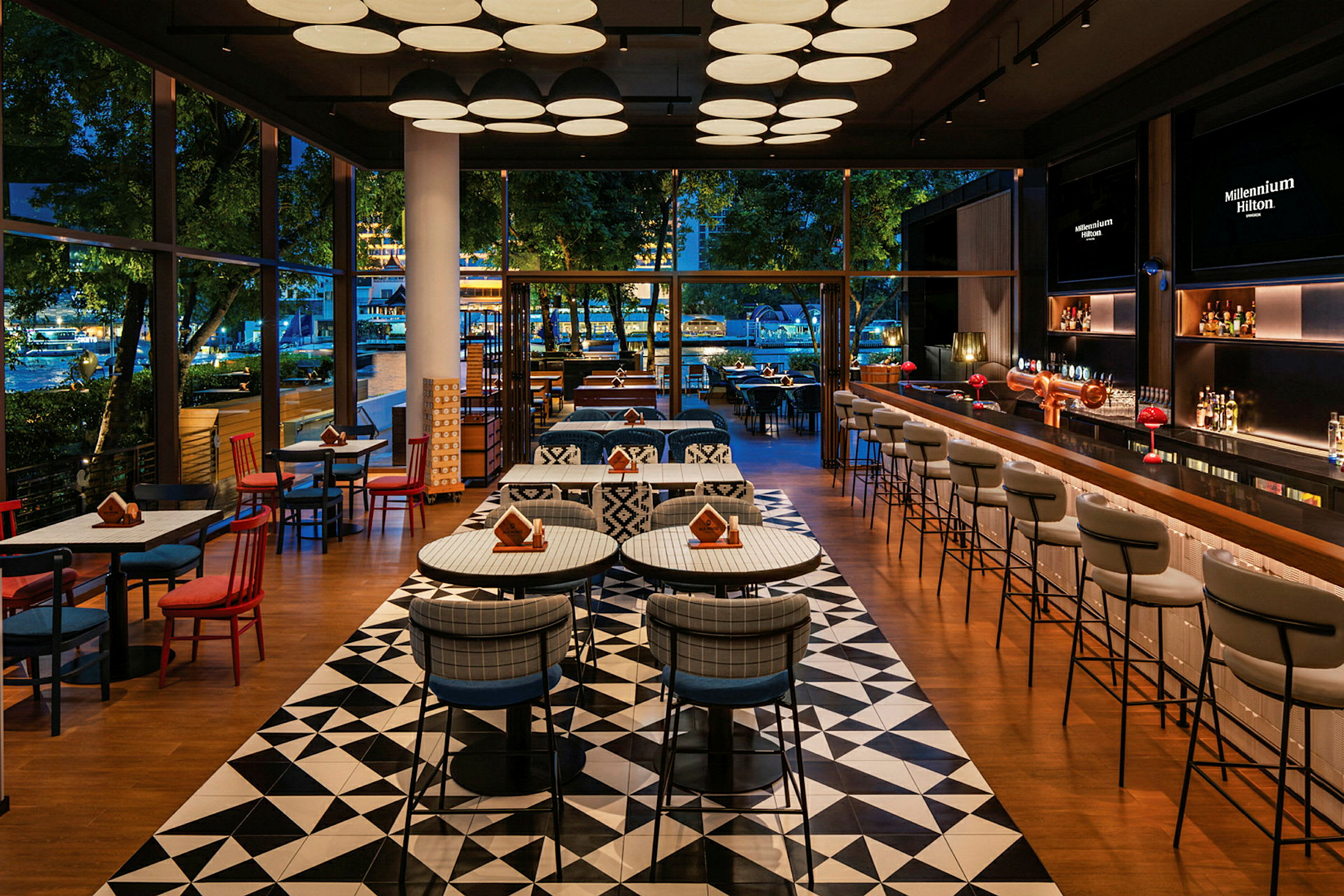 Et elegant restaurantområde med moderne møbler, mønstret gulv og store glasvinduer, der viser udsigt til et udendørsområde med træer. (AI-genereret tekst)