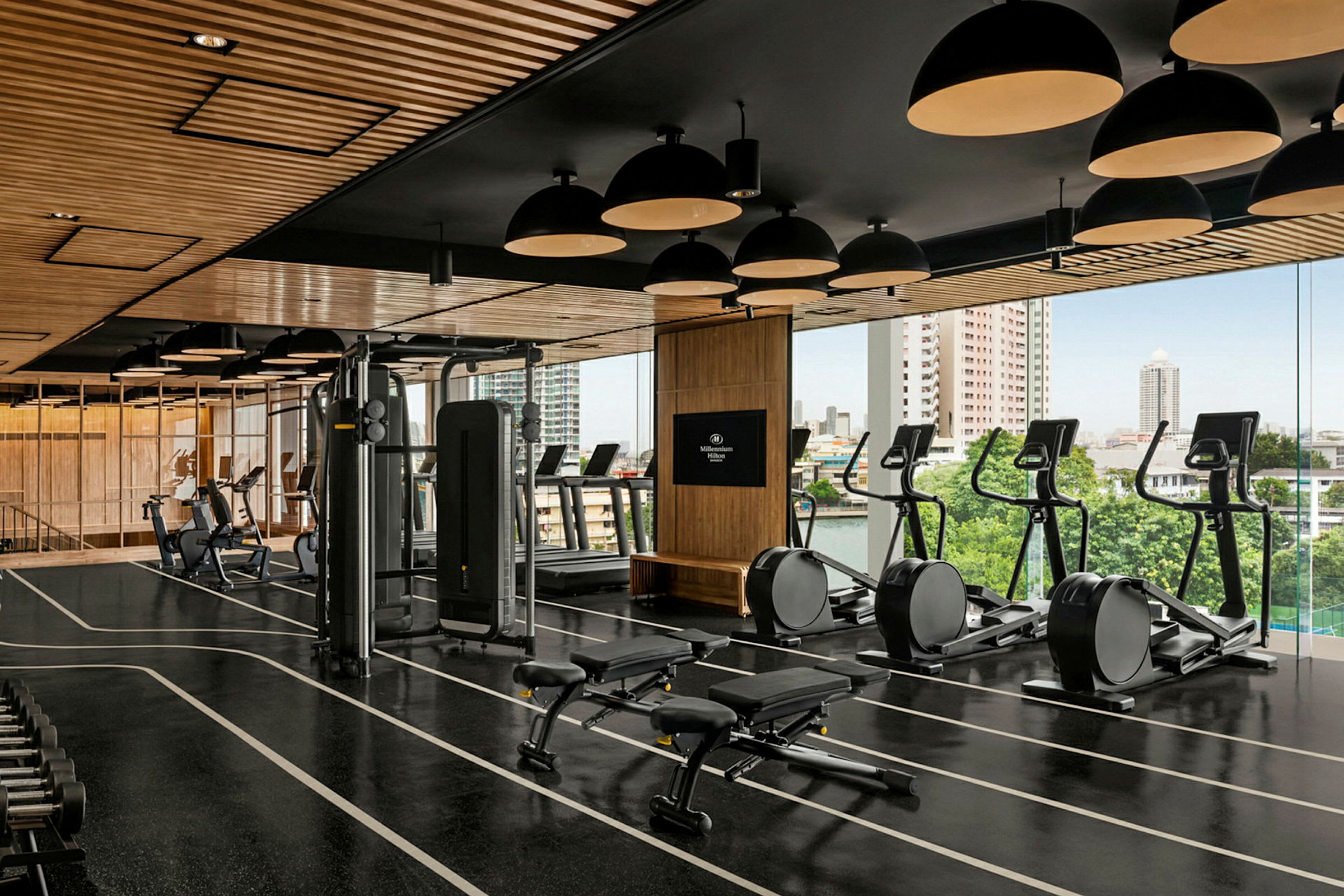 Et veludstyret fitnesscenter med træningsmaskiner, frie vægte og bænke. Lokalet er lyst med store vinduer, der giver udsigt over byen. (AI-genereret tekst)