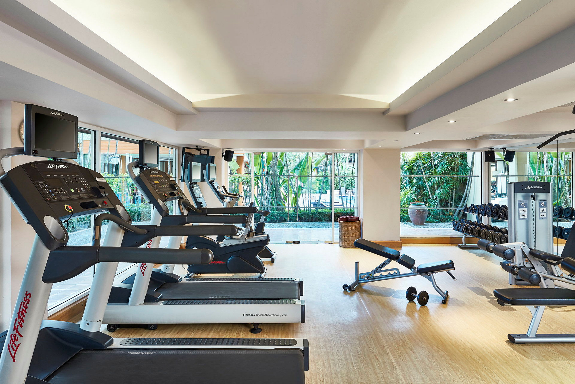 Et fitnesscenter med træningsmaskiner og vægte. Vinduer vender ud mod en grøn gårdhave. (AI-genereret tekst)
