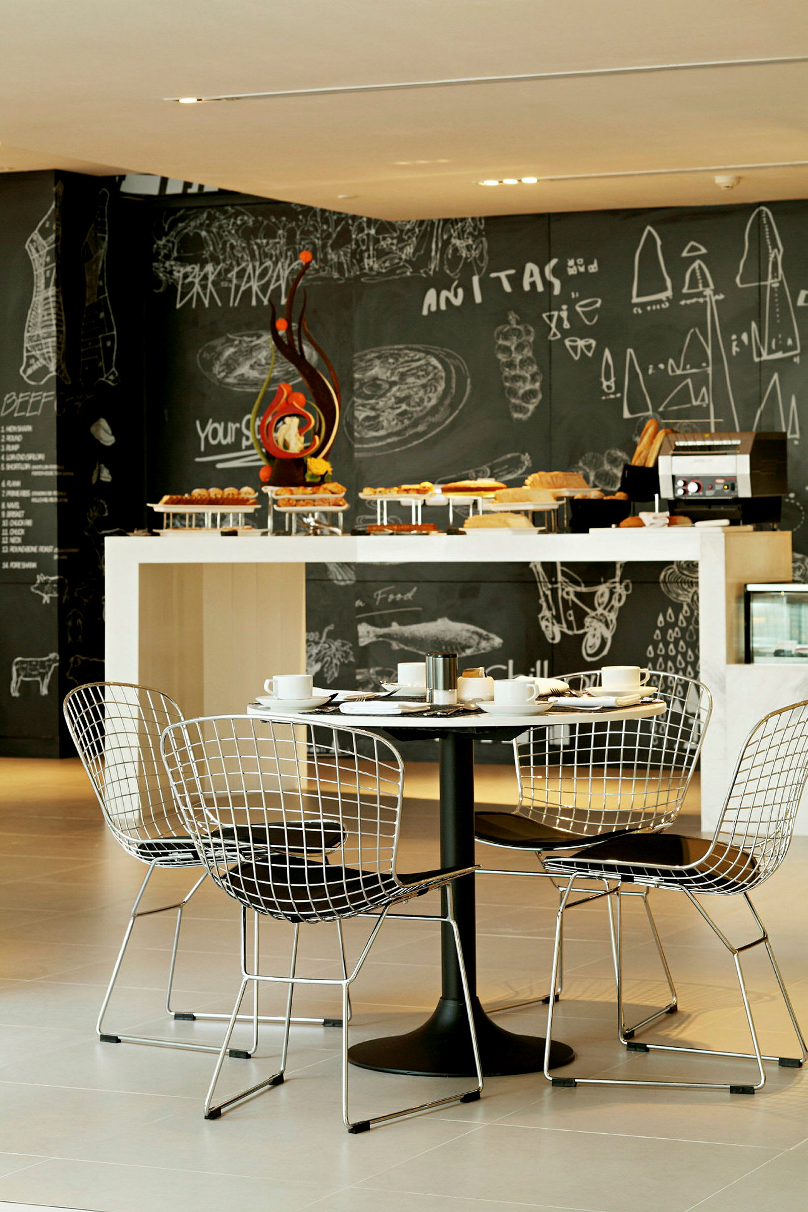 Et moderne restaurantinteriør med et bord og fire stole i forgrunden samt en buffet med forskellige retter foran en sort tavle med tegninger og skrift. (AI-genereret tekst)