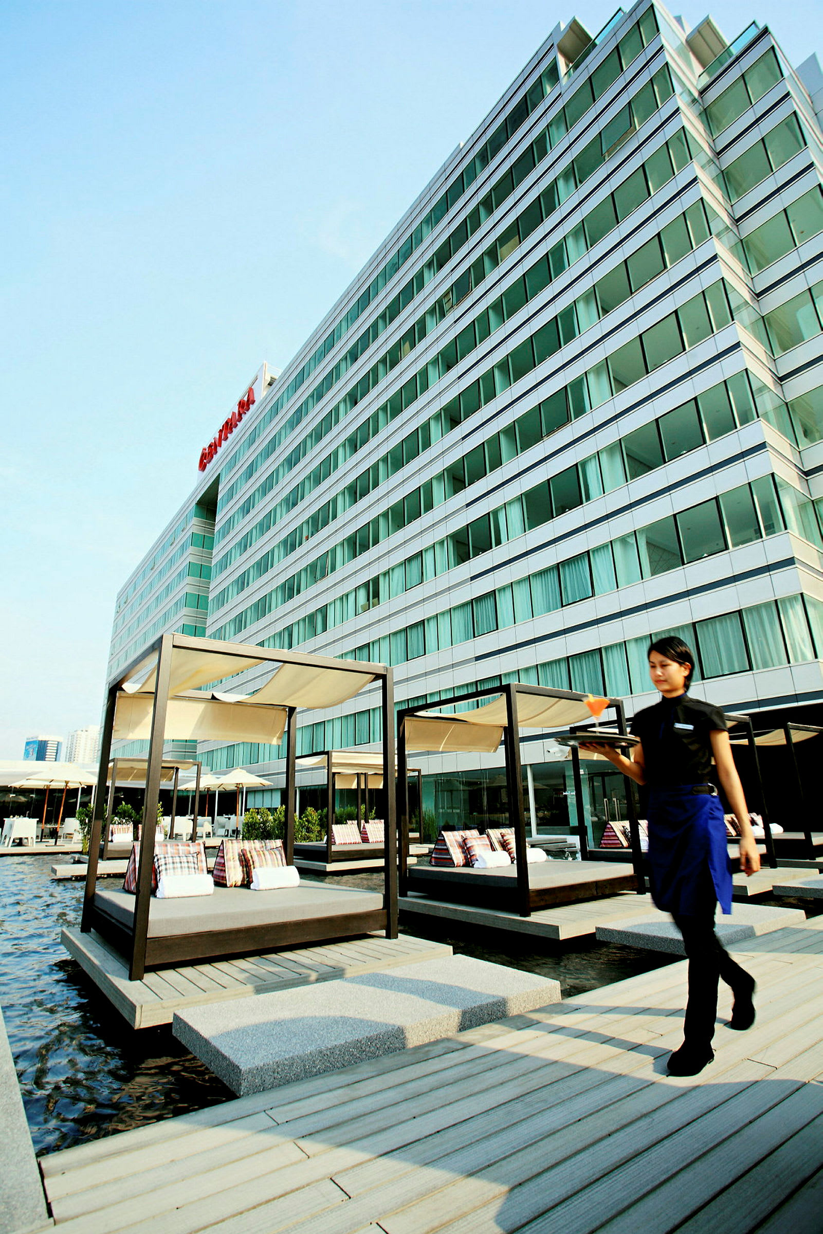 Et moderne hotel med glasfacade og solvogne ved en vandoverflade. En person går med en serveringsbakke. (AI-genereret tekst)