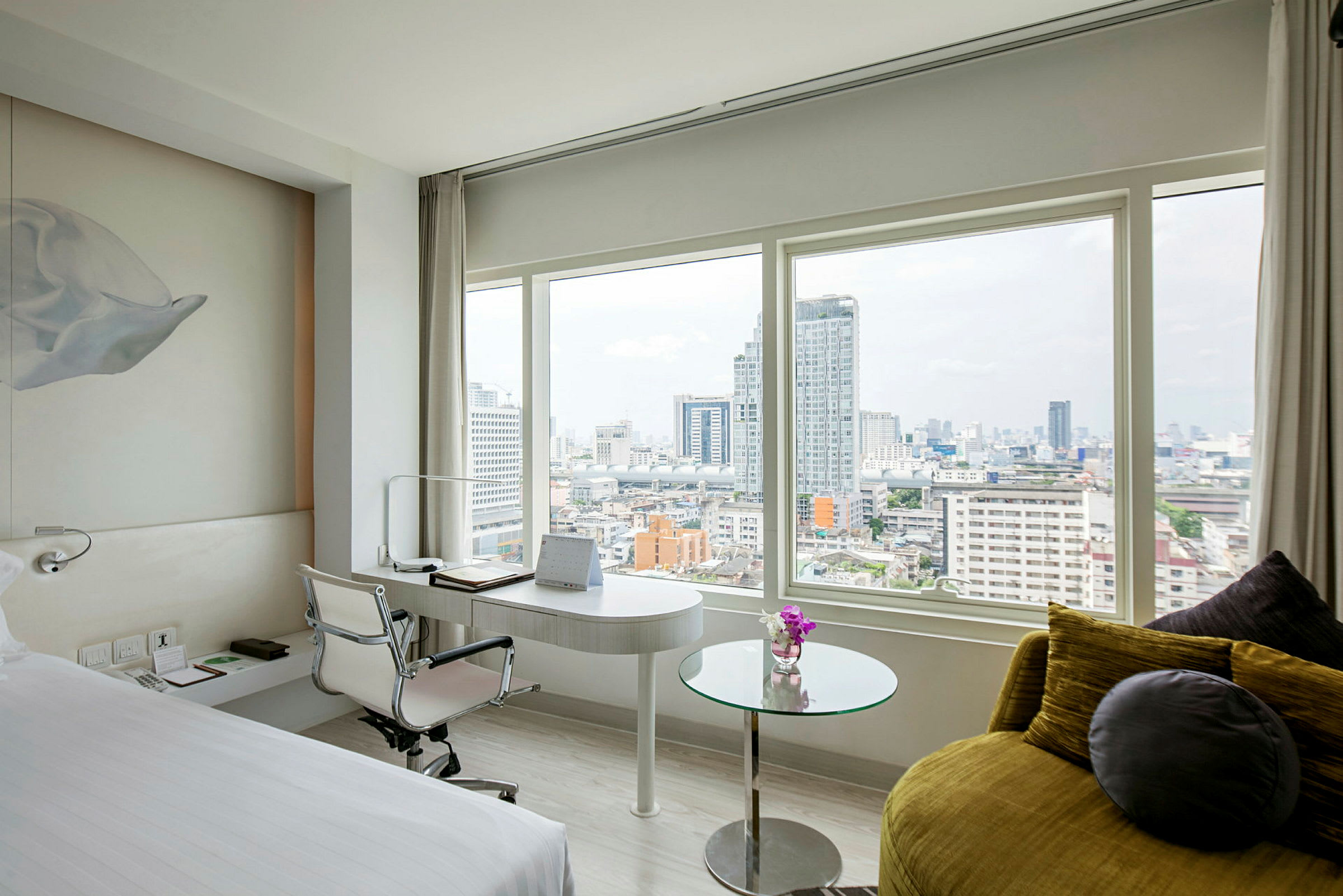 Et moderne hotelværelse med en stor seng, et skrivebord og et glasbord med udsigt over byens skyline gennem store vinduer. (AI-genereret tekst)