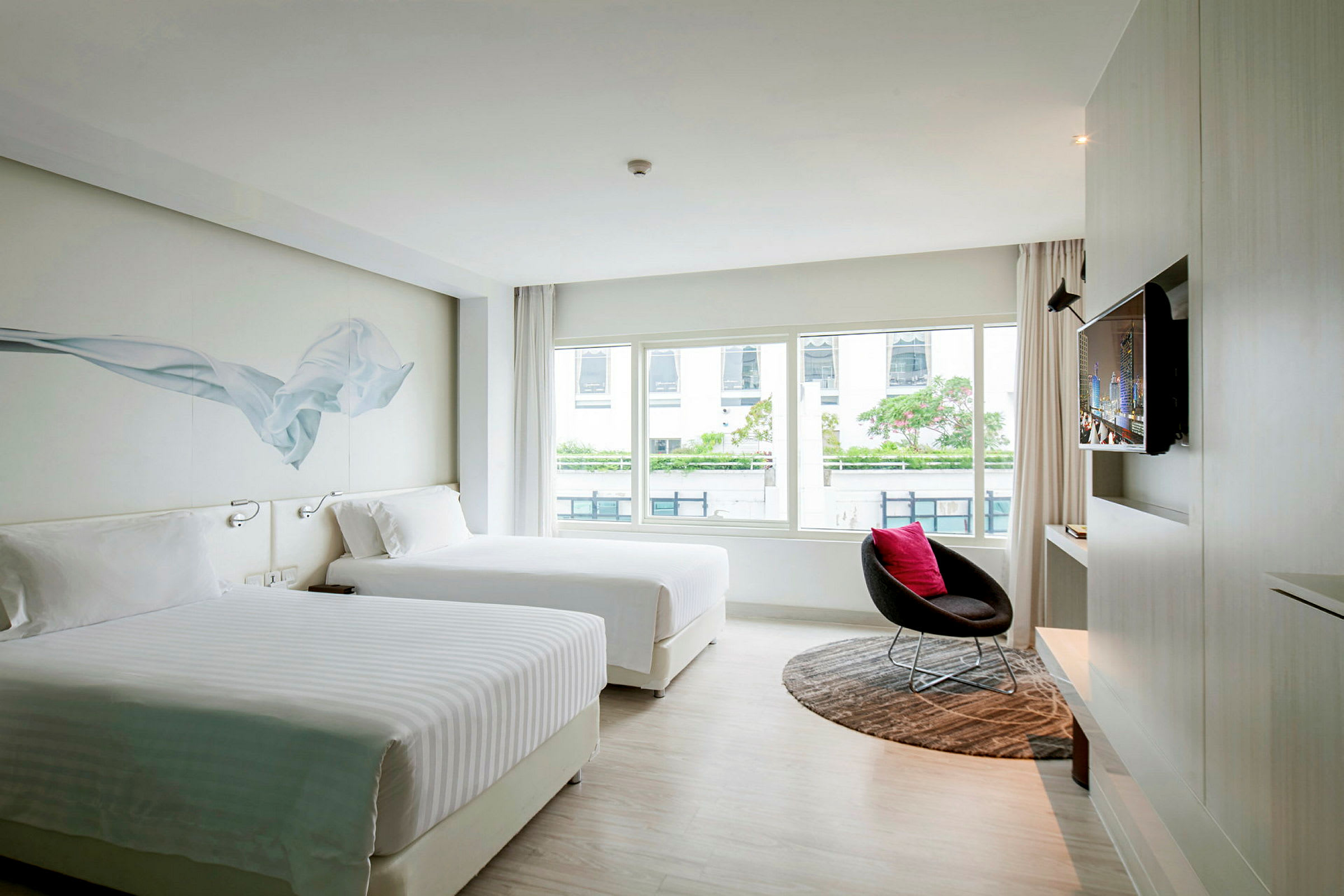 Et moderne hotelværelse med to store senge, en stor vinduesvæg med udsigt, et fjernsyn på væggen og en stol med et rundt tæppe. (AI-genereret tekst)